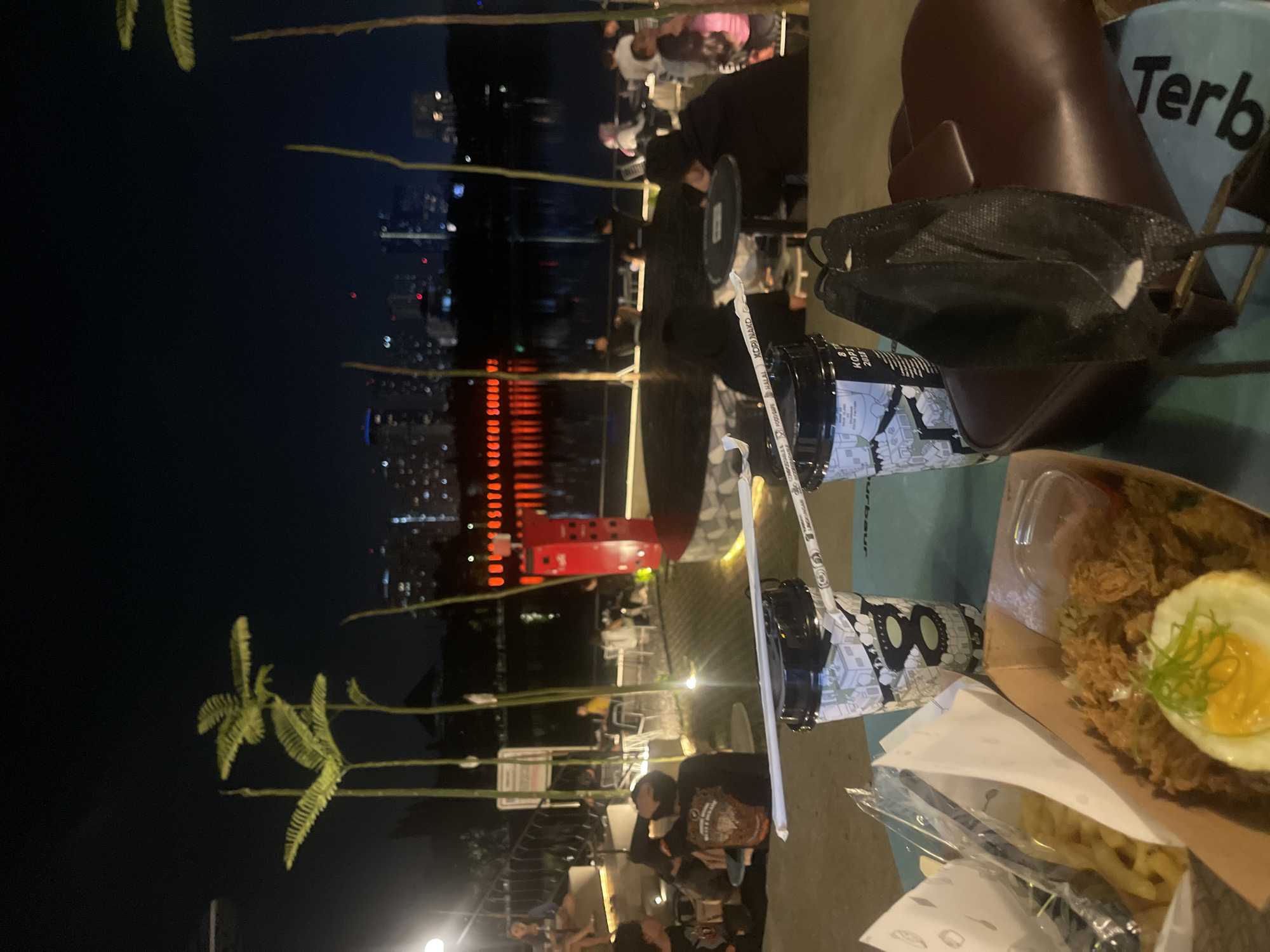 The image captures an outdoor nighttime scene at what appears to be a waterfront dining or leisure area in Surabaya, Indonesia. In the foreground, a turquoise table holds two disposable coffee cups with straws, a brown leather handbag, and a black face mask. To the left, a cardboard container holds fried rice topped with a fried egg and garnished with green onions, next to a container of french fries.
In the mid-ground, several people are seated at tables, some with umbrellas. Tall, thin stalks of vegetation, possibly bamboo, are visible throughout the scene, with fern-like foliage at the top of some. Behind the seated patrons, a body of water reflects the lights of the city skyline. A prominent red structure with illuminated red horizontal lights is situated along the waterfront, casting a vibrant glow on the water.
The background features a cityscape with numerous buildings, some of which are illuminated. The overall atmosphere is that of a casual evening gathering, likely at a restaurant or cafe by a lake or river in Surabaya, illuminated by artificial lighting.