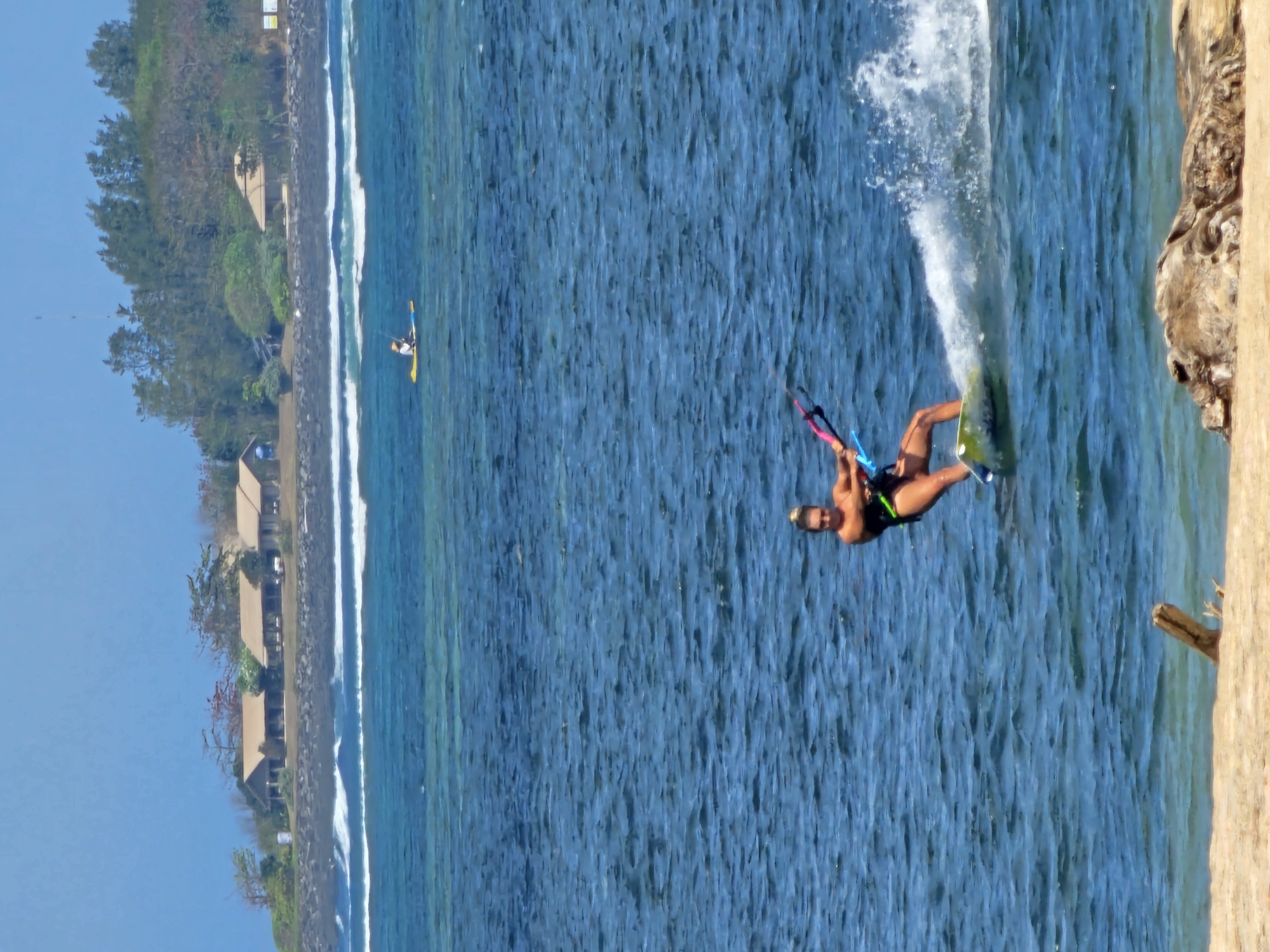 A kite surfer is shown in the ocean, performing a maneuver that kicks up a spray of water. The surfer is wearing a black swimsuit and appears to be in motion on a colorful kiteboard. In the background, a person is visible on a yellow paddleboard in the distance. Further in the background, a coastline with several buildings and trees is visible under a clear blue sky. The scene appears to be a tropical or coastal environment, likely located in Denpasar, Indonesia, given the presence of the ocean and beach. The foreground shows a sandy beach with a piece of driftwood.