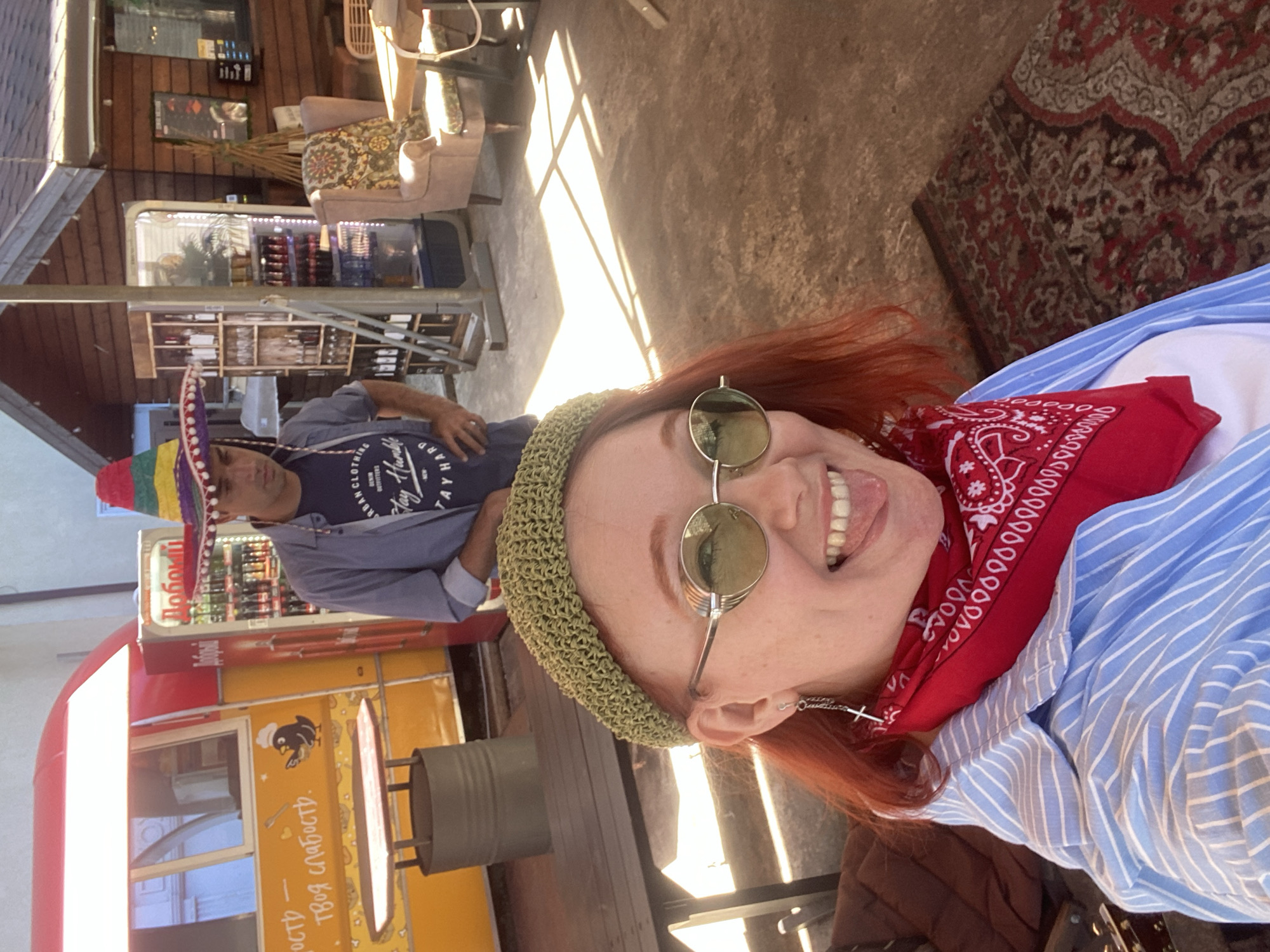 The image depicts a vibrant outdoor scene, likely a cafe or market setting in Sochi, Russia, under bright daylight. In the foreground, a woman with red hair, wearing a green crocheted headband, round sunglasses, and a red bandana, smiles playfully at the camera with her tongue extended. She is dressed in a blue and white striped shirt and sports a silver cross earring.
Behind her, partially obscured, a man in a colorful Mexican sombrero and a dark blue t-shirt stands near a wooden display. His shirt bears the text "TAY HUMBLE TAY HARD" and "DENIM URBAN CLOTHING." The background features a red and yellow food truck with a cartoon penguin chef logo and Cyrillic text reading "Добоми" and "— твоя слабость." A grey metal barrel is positioned beside the food truck.
The setting also includes wooden shelving stocked with bottles, a refrigerator displaying various items, and a seating area furnished with patterned armchairs and a decorative patterned rug on the ground. Strong sunlight casts distinct shadows, indicating a clear, sunny day. The overall mood is casual and cheerful, suggesting a leisurely outdoor activity.