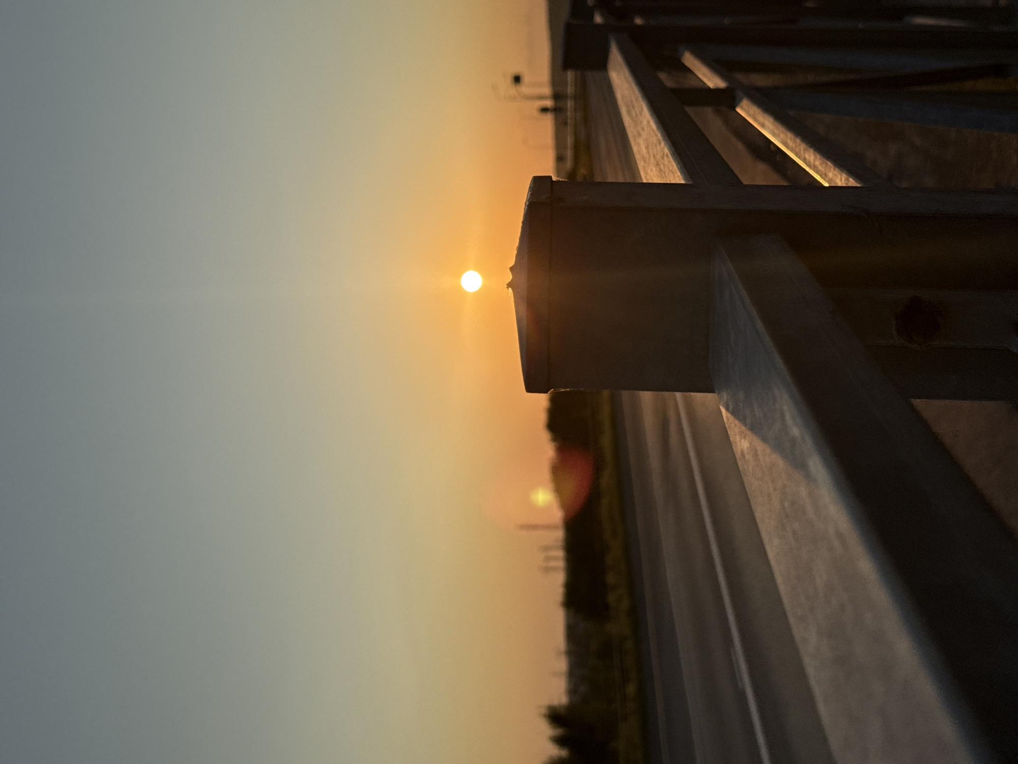 The image shows a sunset over a highway with a metal railing in the foreground. The sun is a bright orb in the sky, casting a warm glow over the scene. The highway is empty, with no cars visible. Trees line the side of the road, and in the distance, there are some industrial structures. The sky is a gradient of orange and blue, with some lens flare from the sun. The railing is made of metal and has a weathered appearance. The overall mood of the image is peaceful and serene.
