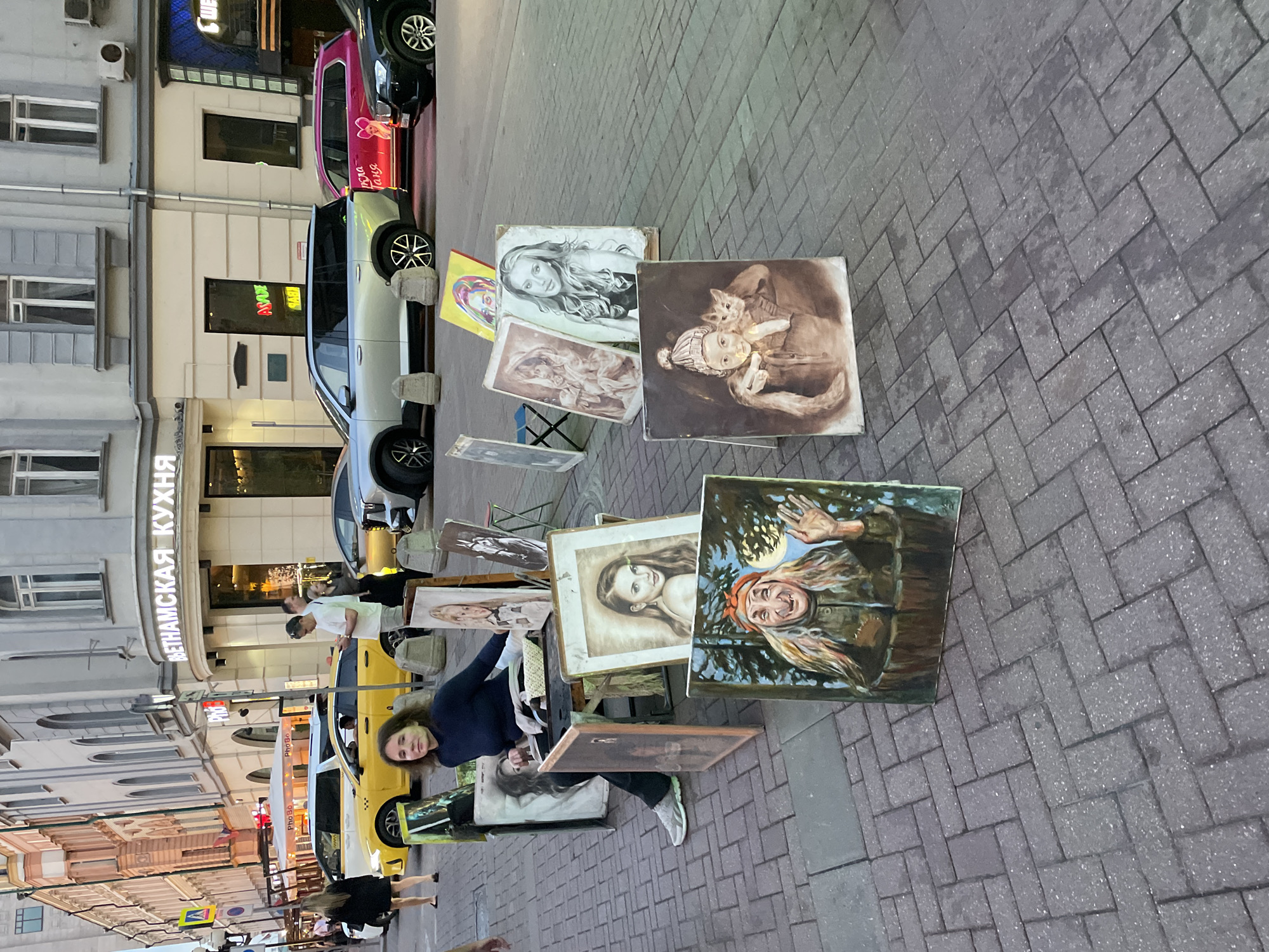 The image shows a street scene in Moscow, Russia, with artwork displayed for sale on the sidewalk. In the background is a building with a sign in Russian that includes the words "Вьетнамская Кухня," meaning Vietnamese cuisine. Various paintings are propped up on small easels or leaning against each other, including portraits of women and other figures. A woman with dark hair is seen standing near the paintings. Passersby can be seen walking on the street, and there are cars parked along the curb. The sidewalk is paved with grey bricks. The scene gives the impression of an outdoor art market in a bustling city environment.