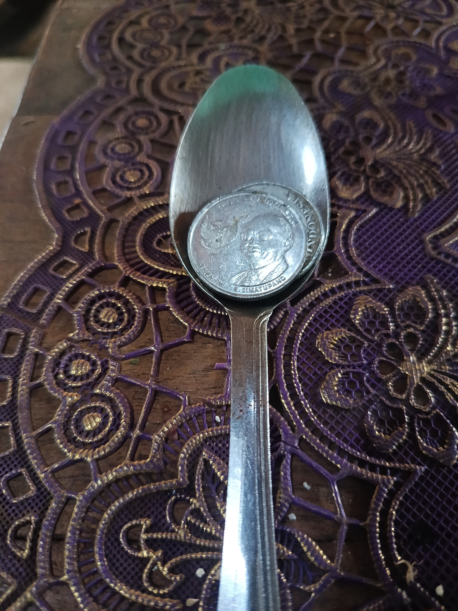 An overhead shot captures a metal spoon resting on a decorative placemat. The spoon is positioned vertically down the center of the frame, with its bowl containing two silver coins. The placemat, a rich purple fabric adorned with intricate gold patterns, lies on a wooden surface. The image appears to be a close-up, with the spoon and placemat filling most of the frame. No people are visible in the image, and no specific location in Karangmojo, Indonesia, can be identified from the visual information provided. The lighting is even, suggesting an indoor setting.