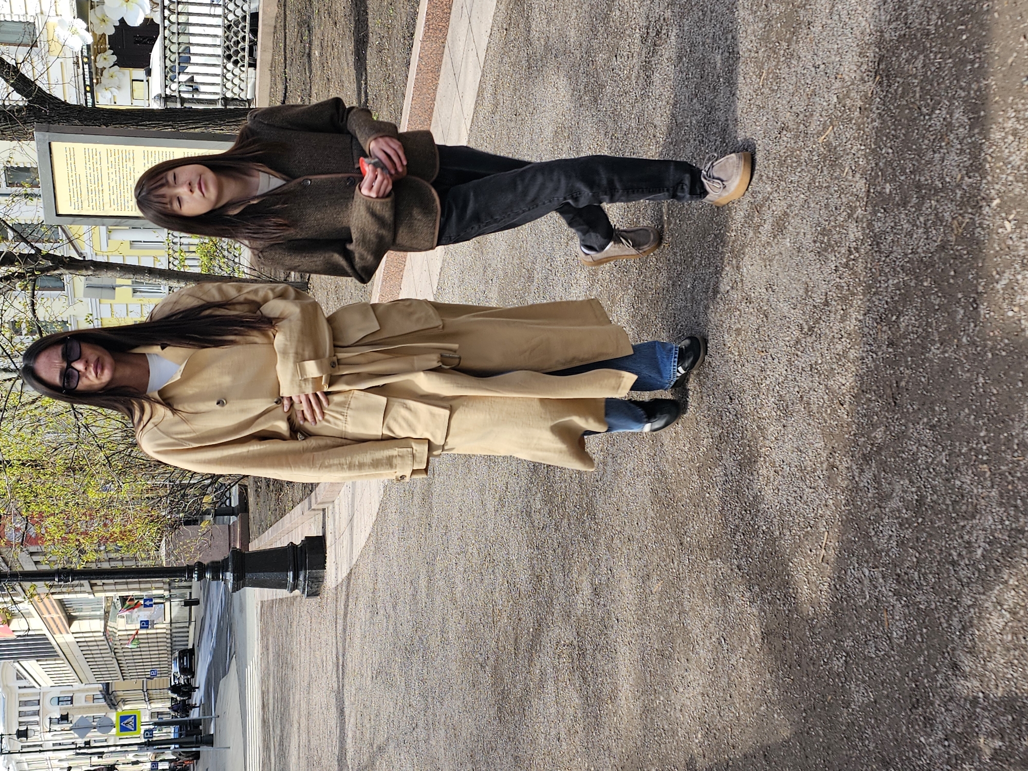 The image depicts two women standing side-by-side on a paved sidewalk. The woman on the right wears a brown, tweed-like jacket, dark jeans, and tan shoes. She is holding a small object in her hands. The woman on the left is wearing a tan trench coat, blue jeans, and black shoes. She is also wearing sunglasses.
In the background, there is a street with buildings, trees, and traffic lights. There is a sign or monument behind the women. The location appears to be in Moscow, Russia.