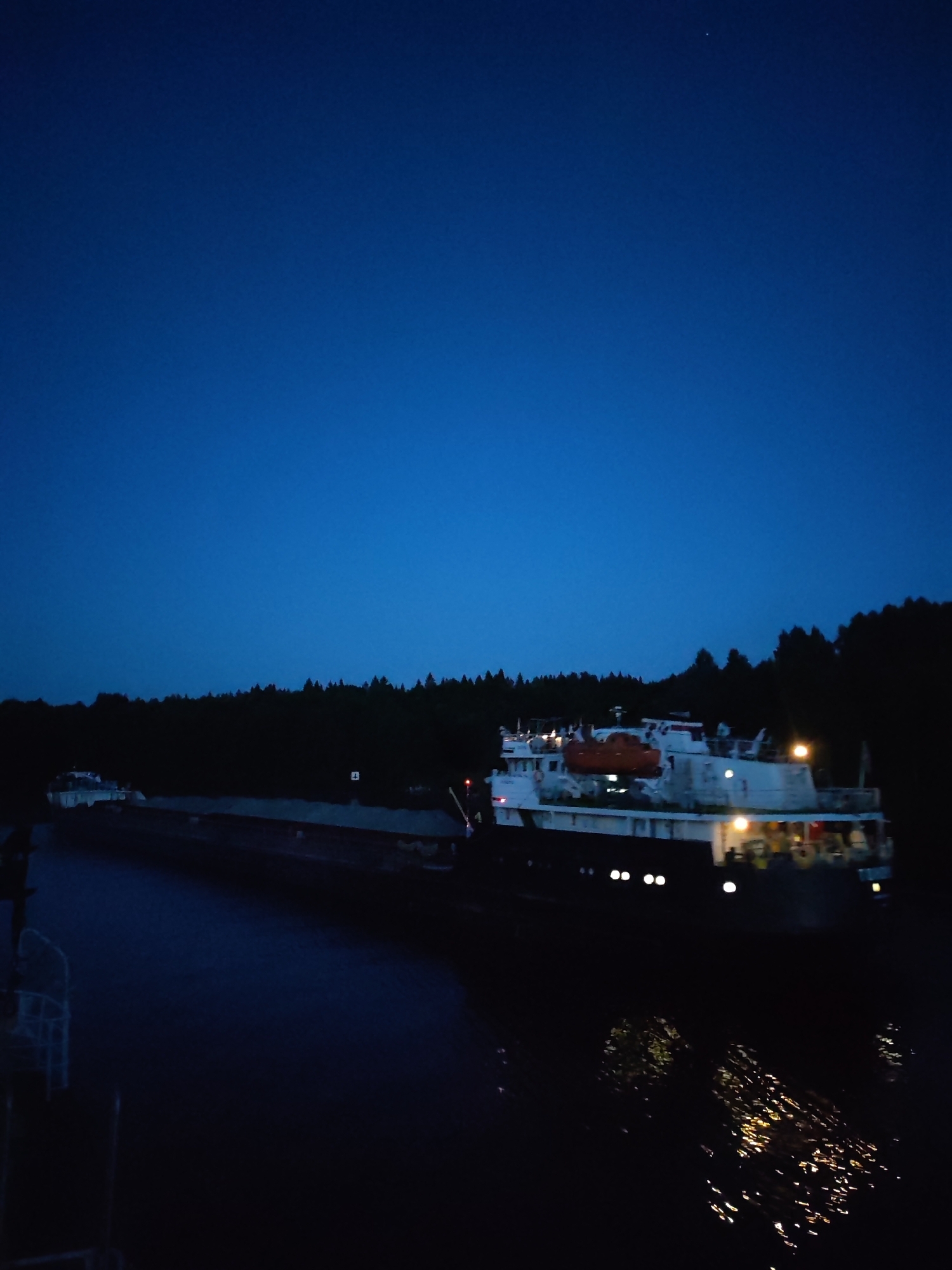 The image depicts a nighttime scene in Volokov Most, Russia, with a clear, dark blue sky overhead, indicating twilight or early night. A vast expanse of water occupies the lower half of the frame, reflecting the lights of the vessels and the distant tree line. A long, dark barge is positioned in the foreground, its structure barely discernible in the dim light. Behind it, a white vessel with illuminated windows and an orange lifeboat is visible. The barge and the vessel appear to be on a river or canal, as evidenced by the calm water. The background is dominated by a dark, dense forest line, silhouetted against the sky, with trees suggesting a natural, possibly rural, setting. The overall atmosphere is serene and quiet.