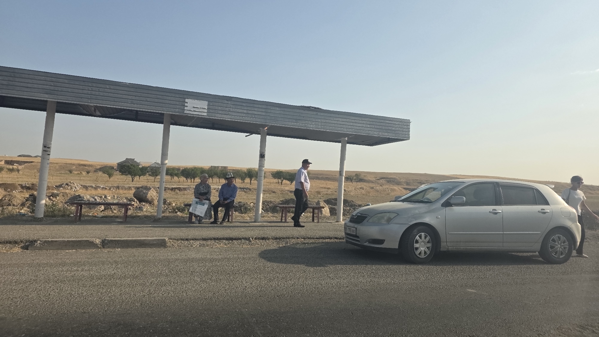 The image depicts a bus stop in Baş-Bulak, Kyrgyzstan. A silver hatchback car is parked on the right side of the frame, facing left. A woman in a white t-shirt and dark pants stands next to the car. To the left of the car is a bus stop shelter with a metal roof supported by white pillars. Three individuals are seated on a bench under the shelter: two elderly women and a man wearing a traditional Kyrgyz hat. Another man in a white shirt and dark pants stands in the shelter. The environment is arid with dry grass and scattered trees visible in the background, under a clear blue sky.