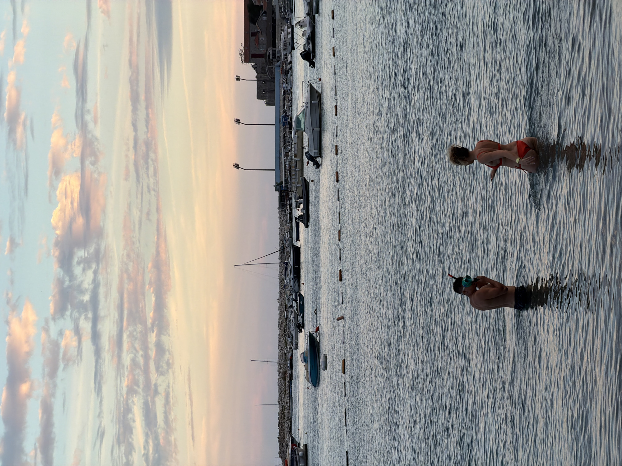 The image depicts a serene scene at sea, with two individuals wading in the water. In the foreground, a man wearing a snorkel mask stands facing away from the viewer, with his hands near his mouth. Beside him, a woman in a red bikini is seen from the back, with her left arm extended slightly. They appear to be engaged in conversation.
The water is calm, with gentle ripples reflecting the soft hues of the sky. In the background, a harbor is filled with numerous boats of various sizes, anchored near a stone breakwater. Beyond the harbor, a cityscape is visible, characterized by buildings with reddish-brown roofs, and tall, modern light poles. The sky is painted with a gradient of colors, from soft blues to warm pinks and oranges, suggesting a sunset or sunrise.
Based on the distinctive architecture and the specific configuration of the harbor and surrounding buildings, the scene is identifiable as the coast of Budva, Montenegro. The overall atmosphere is tranquil and idyllic, capturing a moment of leisure by the Adriatic Sea.