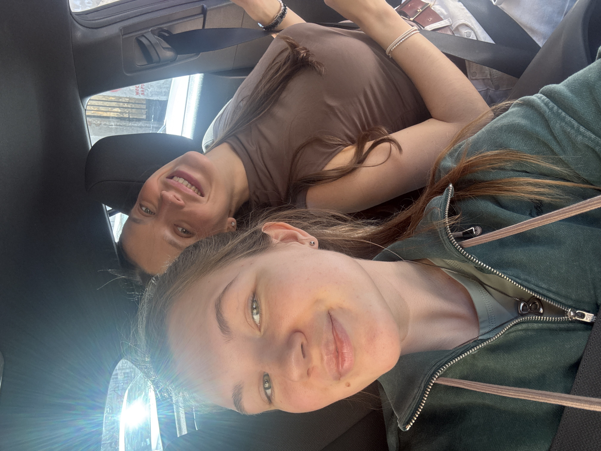 Two females are inside a vehicle. The younger female wears a green zip-up jacket. The older female wears a brown shirt. Both are smiling. They are wearing seatbelts. The location is Belgrade, Serbia, as evidenced by a Cyrillic script sign visible through the window.