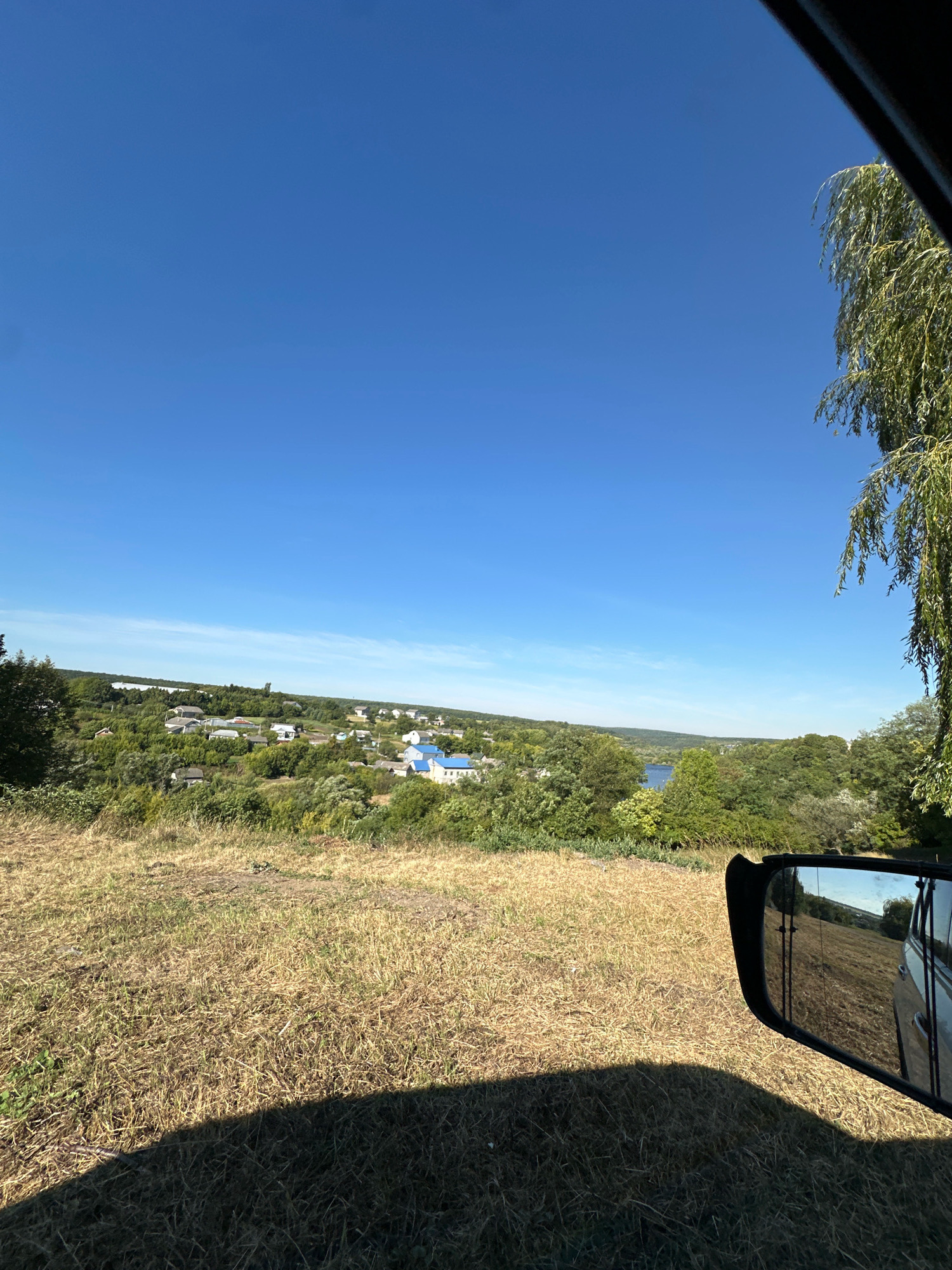 An outdoor, eye-level, wide shot captures a sunlit, rural landscape in Makiv, Ukraine. The scene is viewed from within a vehicle, with a side-view mirror and a portion of the car’s interior frame in the foreground. The mirror reflects a grassy hillside and a blue sky. A large, leafy tree hangs into the frame from the upper right. Beyond the foreground, a village is nestled amongst rolling, green hills and dense vegetation. The village is characterized by numerous houses, many with blue rooftops, scattered across the landscape. A body of water, likely a river or lake, is visible in the distance. The sky is a clear, vibrant blue. The ground in the immediate foreground is covered in dry, yellowed grass, casting a long shadow from the vehicle.