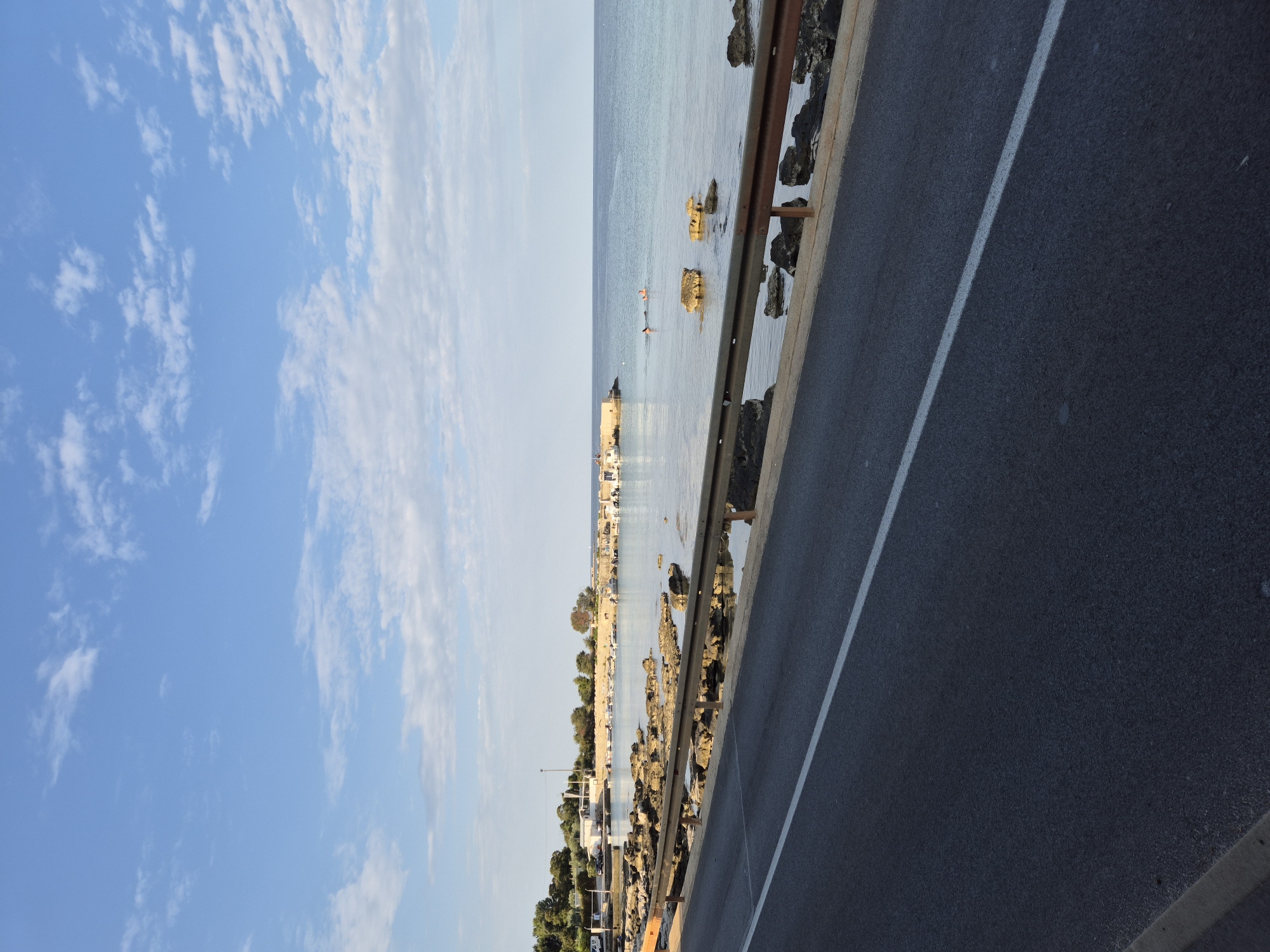 The image depicts a coastal scene in Zambratija, Croatia, with a focus on the road and the sea. In the foreground, a dark asphalt road with a white dividing line runs diagonally across the frame. A metallic guardrail separates the road from the rocky shoreline. The shoreline is characterized by scattered rocks and boulders, with the calm, blue sea beyond. Several small boats are moored in the distance, near a pier or breakwater. A few people are visible in the water, swimming or wading. The sky above is a clear blue with scattered white clouds. The overall impression is one of a serene, seaside location during the daytime.