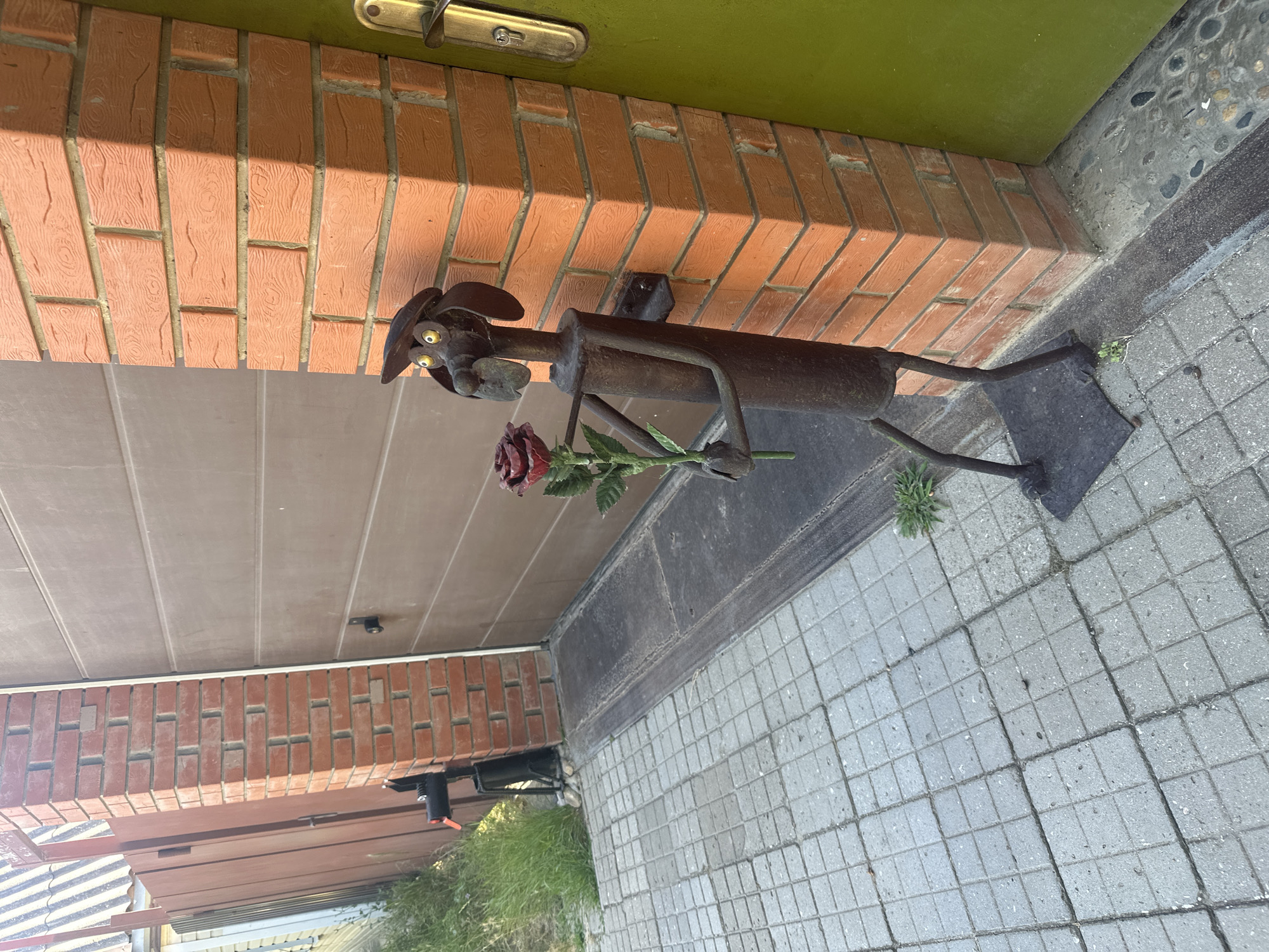 A metal sculpture of a dog holding a red rose stands on a paved area next to a brick pillar and a garage door. The dog sculpture is made of dark metal and has a hat, big eyes, and a snout. The rose appears to be also made of metal, with red petals and a green stem and leaves. The brick pillar is made of orange-red bricks. A brown garage door is visible behind the pillar. To the left of the pillar, a green door is slightly ajar. The ground is paved with square grey tiles. In the background, there is grass and a wooden fence. This scene is located in the city of Temryuk, Russia.
