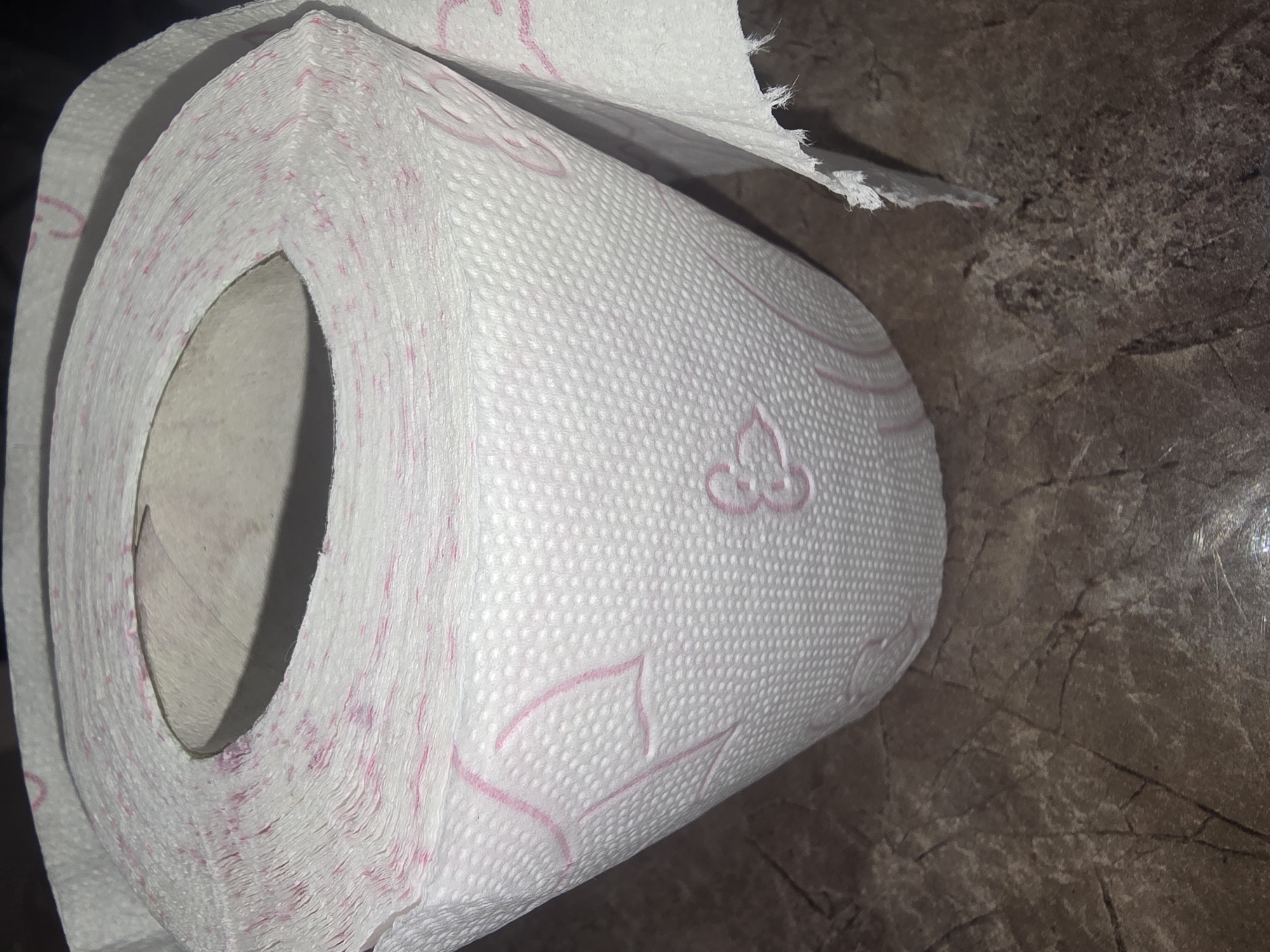 A close-up, low-angle shot shows a roll of toilet paper with a pink pattern on a mottled brown surface. The toilet paper is white with a textured surface and features a repeating pink floral or heart-like design and some scattered pink dots. The roll is partially unspooled, revealing layers of perforated paper. The background appears to be a countertop or table with a marbled or stone-like texture. There are no people or animals present in the image. The image does not provide any information about its location in Ulan-Ude, Russia.
