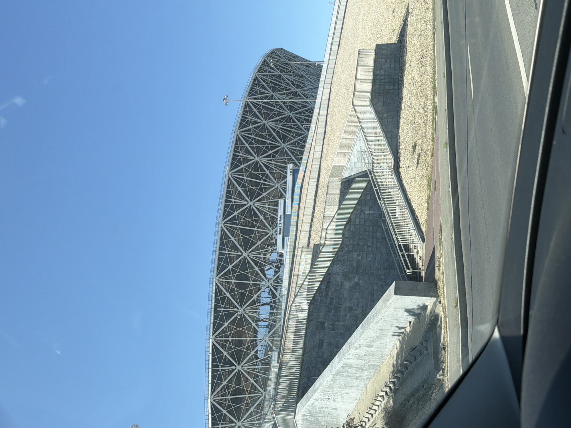 The image shows a view of Volgograd Arena in Volgograd, Russia, on a sunny day. The stadium's distinctive geodesic dome roof structure is visible against a clear blue sky. In the foreground, there are concrete stairs and a walkway leading up to the stadium, with metal railings. The area surrounding the stadium appears to be covered in gravel and some sparse vegetation. A road is visible in the bottom part of the image, suggesting the perspective is from a vehicle. There are no people or vehicles clearly identifiable in the scene.