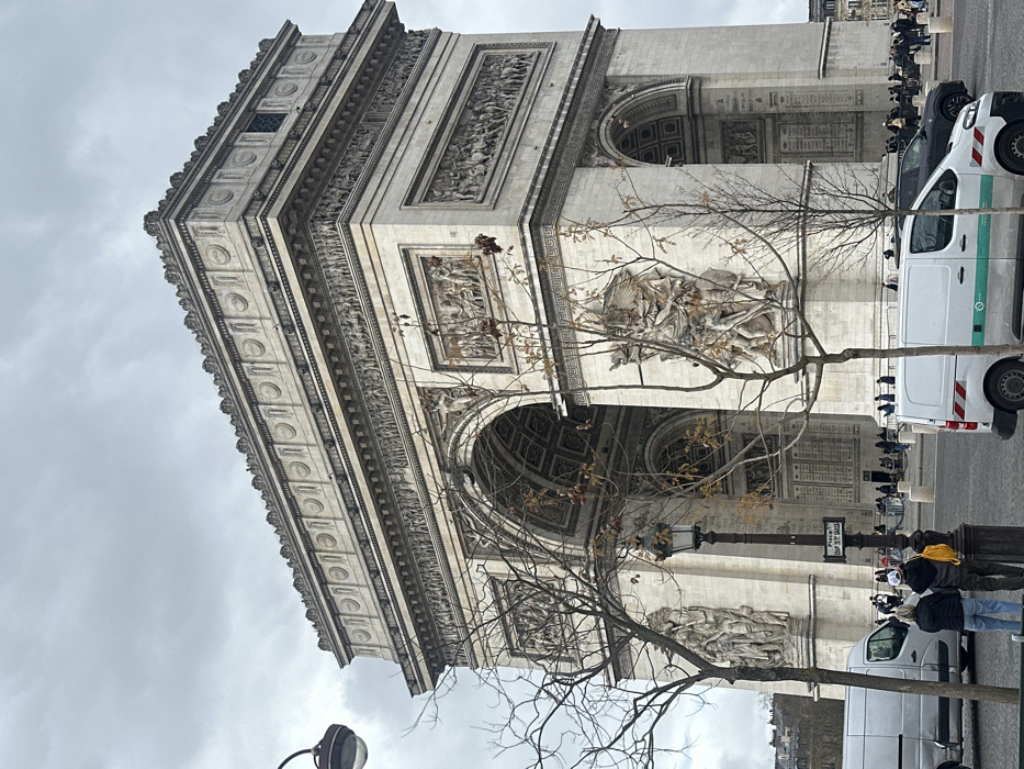 The image depicts the Arc de Triomphe, situated at the western end of the Champs-Élysées on the Place Charles de Gaulle in Paris, France.
The central subject is the monumental Arc de Triomphe, constructed of light-colored stone, dominating the frame under an overcast sky. Its intricate architectural details are visible, including large bas-relief sculptures on its faces, friezes encircling the structure, and ornate cornices along its upper levels. The arch features a large central opening and smaller arches on its sides.
A crowd of people is visible near the base and within the arches of the monument, suggesting public access and tourism. In the foreground, two individuals are seen from behind, facing the Arc. One wears a dark jacket, blue jeans, and tan boots; the other wears a dark jacket, dark pants, and a yellow backpack.
Several vehicles are present. On the left, a silver cargo van is parked. In the foreground-right, a white cargo van with a green stripe and red and white reflective chevron markings on its rear is parked. A dark-colored vehicle is partially visible behind this white van.
Street furniture includes a dark, ornate lamppost with a street sign reading "Place Chmp de Gaulle" (Place Charles de Gaulle) attached, positioned between the two foreground individuals and the Arc. Bare-branched trees with minimal foliage partially obscure parts of the monument. Additional lampposts and bollards are visible along the perimeter of the roadway surrounding the Arc.