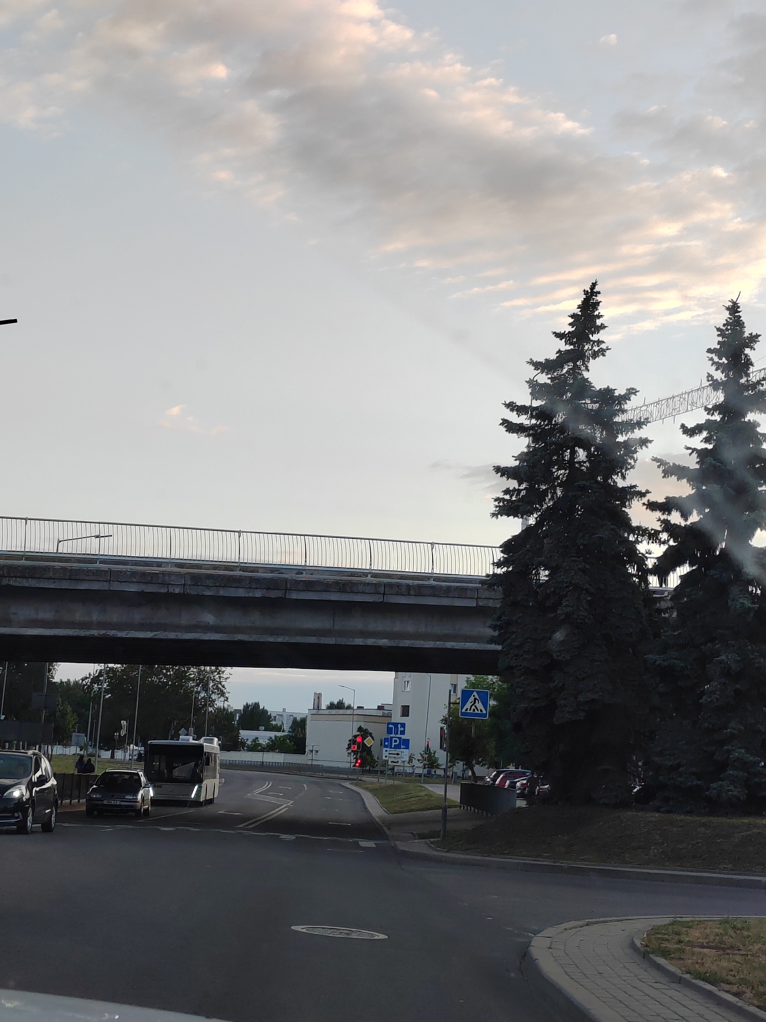 The image captures a street scene in Brest, Belarus, under an overcast sky. In the foreground, a road curves to the right, with a brick-patterned sidewalk. A dark-colored SUV is visible on the left, partially obscured. Further down the road, a silver Volkswagen Golf is parked, and a white bus is in motion. A traffic light shows red.
Overhead, a concrete overpass with a metal railing spans the road. Two tall, dark green evergreen trees stand to the right, partially obscuring a construction crane in the background. Several buildings are visible behind the trees, appearing to be residential or commercial structures. A pedestrian crossing sign is also present. The overall lighting suggests it is either early morning or late evening.