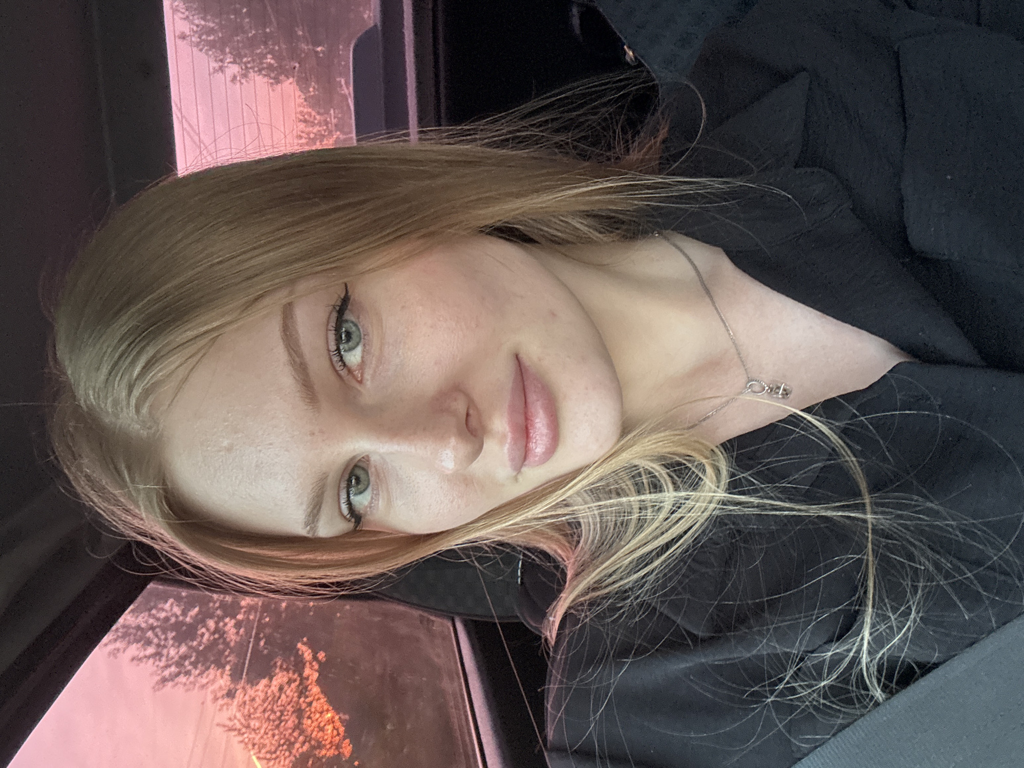 A fair-skinned woman with blonde hair and light-colored eyes is positioned in the foreground, partially obscuring the view outside a car window. Her hair is loose, with strands falling across her dark clothing. She wears a silver necklace. The car window shows an outside scene, tinted with a pink hue, possibly due to the sunset. Vegetation is visible through the window, suggesting a natural environment. Given the location is Кондратово, Russia, this could be a drive through a residential area or on the outskirts of the city.