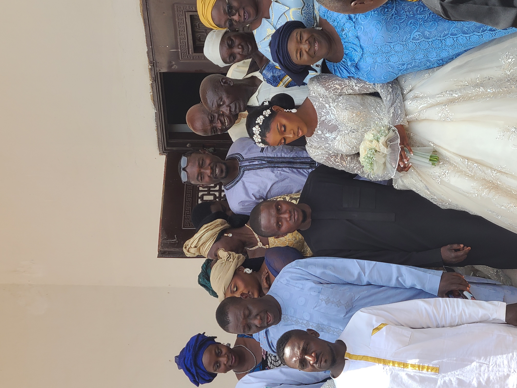 A group of people are posing for a photo in Barakin Lerawi, Nigeria. The focal point is a bride in a white, shimmering wedding dress and holding a bouquet. She is surrounded by men and women, several of whom are wearing traditional African clothing and head coverings. The individuals are smiling and looking toward the camera. An architectural detail is visible behind the group.