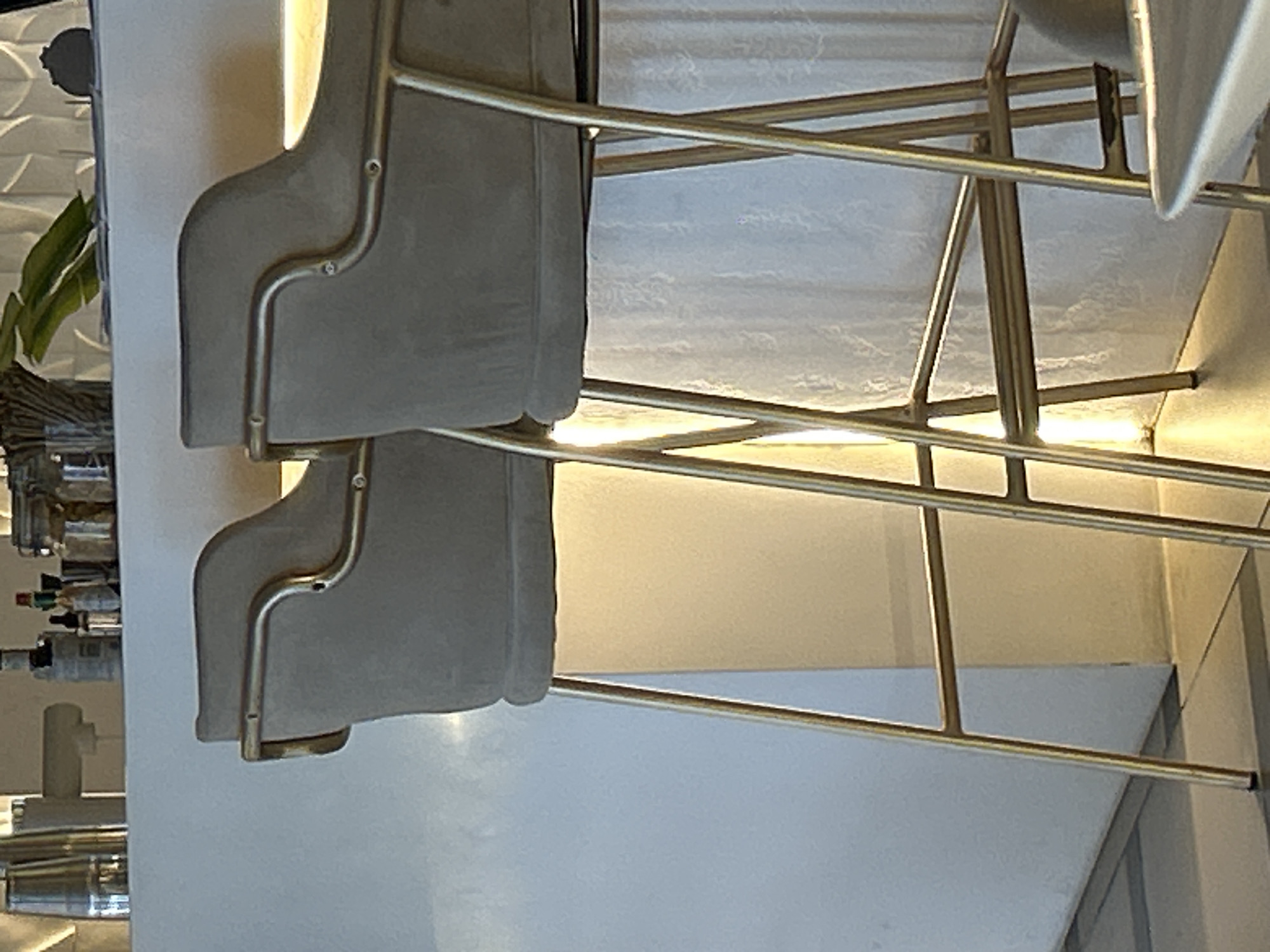 The image depicts two grey upholstered bar stools with gold metal legs, positioned in front of a white counter. The counter has several items on it, including bottles and jars. The stools are illuminated from behind, creating a warm glow. The background features a textured white curtain. The image was taken in North Kuta, Indonesia.