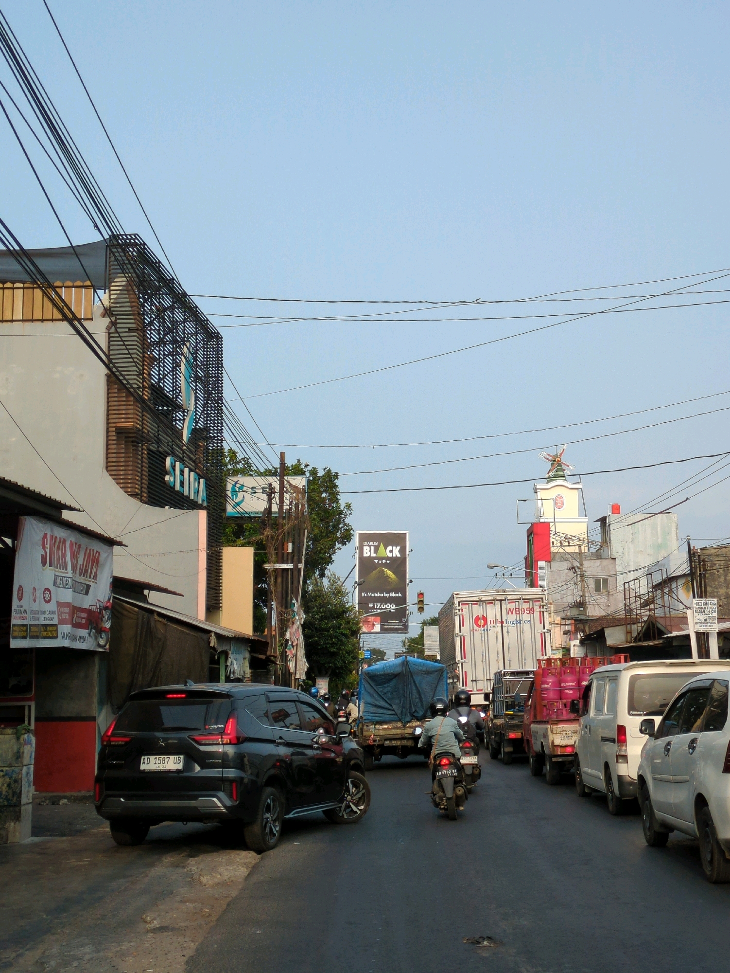 The image captures a street scene in Gentan, Indonesia, during daylight, characterized by moderate traffic and an urban environment. A black SUV, license plate AD 1587 UB, is prominently featured in the foreground on the right side of the road, facing away from the camera. Several other vehicles, including trucks, vans, and motorcycles, are visible further down the street, indicating traffic congestion. Buildings line both sides of the road, some appearing to be commercial establishments with signage. One building on the left has a large sign that reads "SEIRRA". Overhead, a complex network of electrical wires crisscrosses the sky, a common sight in urban Indonesian settings. A billboard advertising "DIARUM BLACK MATCH". The sky is clear and blue, suggesting fair weather. The overall atmosphere is that of a busy, everyday street in a town or city.