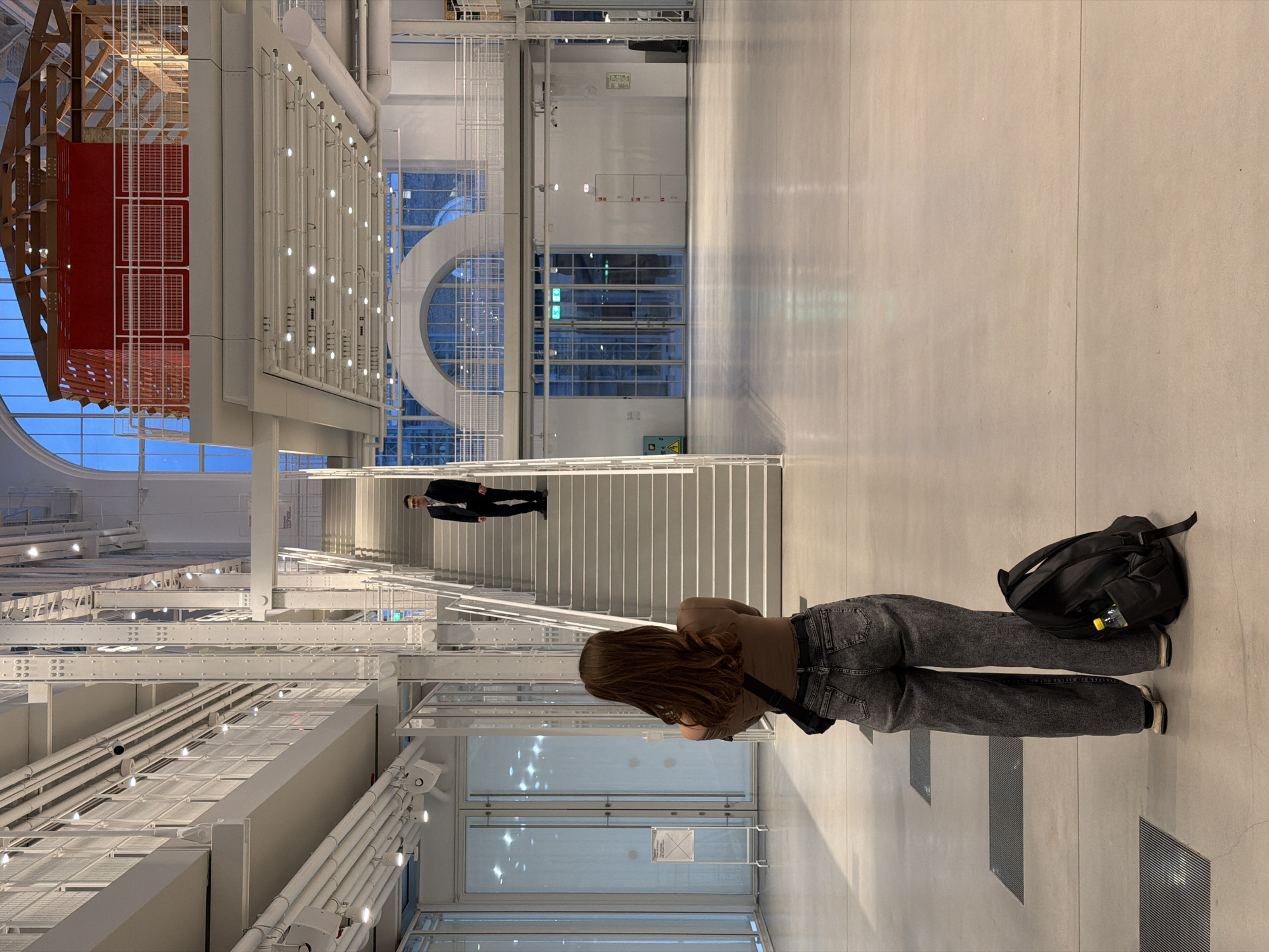 The image shows an interior view of a modern, architectural space. A woman with long brown hair, wearing a brown top and grey jeans, stands with her back to the viewer. A black backpack lies near her feet. Above her, a staircase ascends, and a man in a dark suit walks up the stairs. The architectural style is characterized by white walls, exposed structural elements, and large windows with views to the outside. On the left, a complex structure with wooden and red elements is visible. The environment appears sterile and well-lit. The location is Unknown, Unknown.