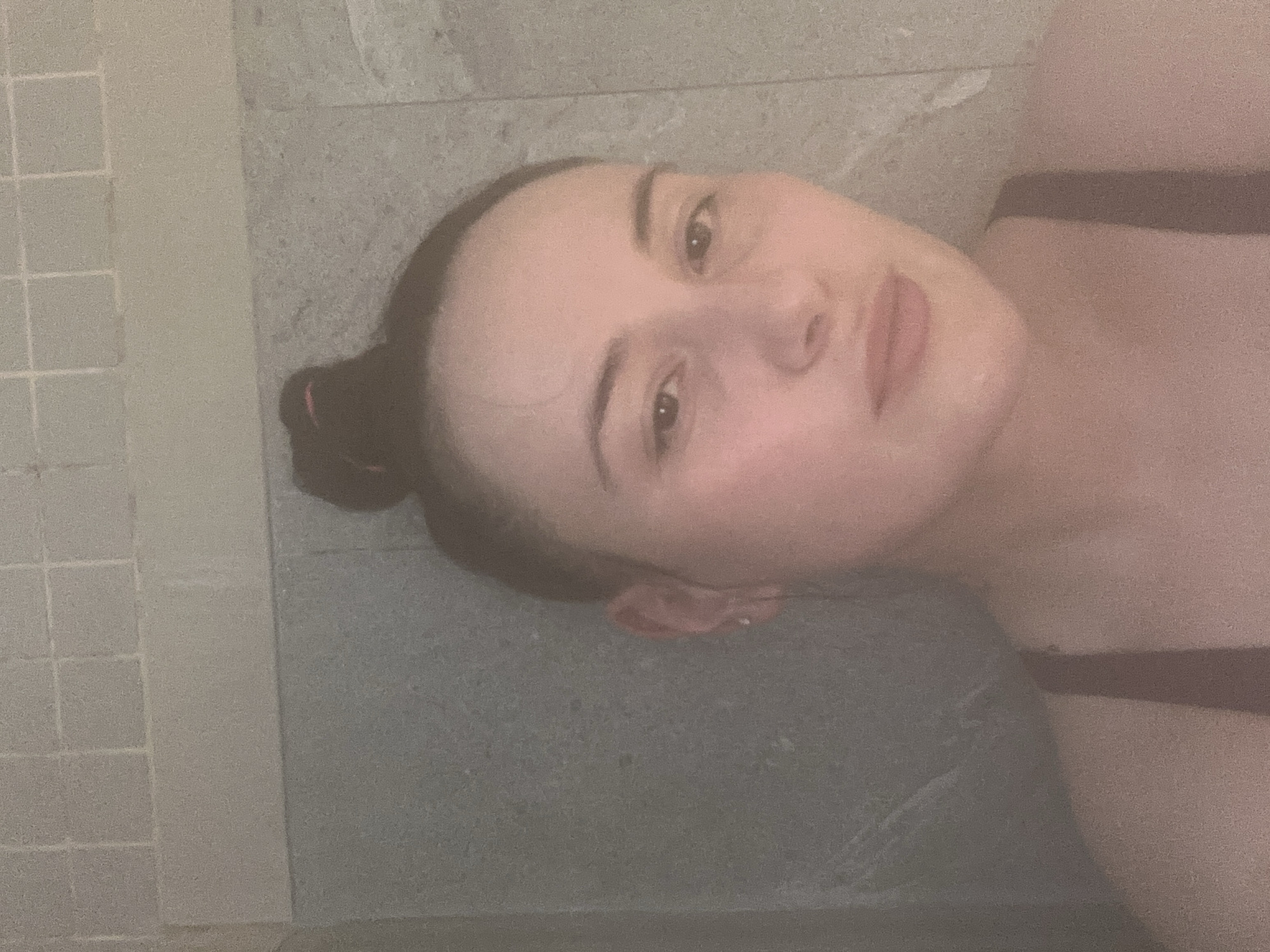 A woman with fair skin and her hair pulled back in a bun is lying on a gray surface. She is wearing a dark-colored top or bathing suit. To her left, a wall is tiled with light gray squares. The scene appears to be a bathroom or spa-like setting. There is no indication of the location being Chaikovsky, Russia.