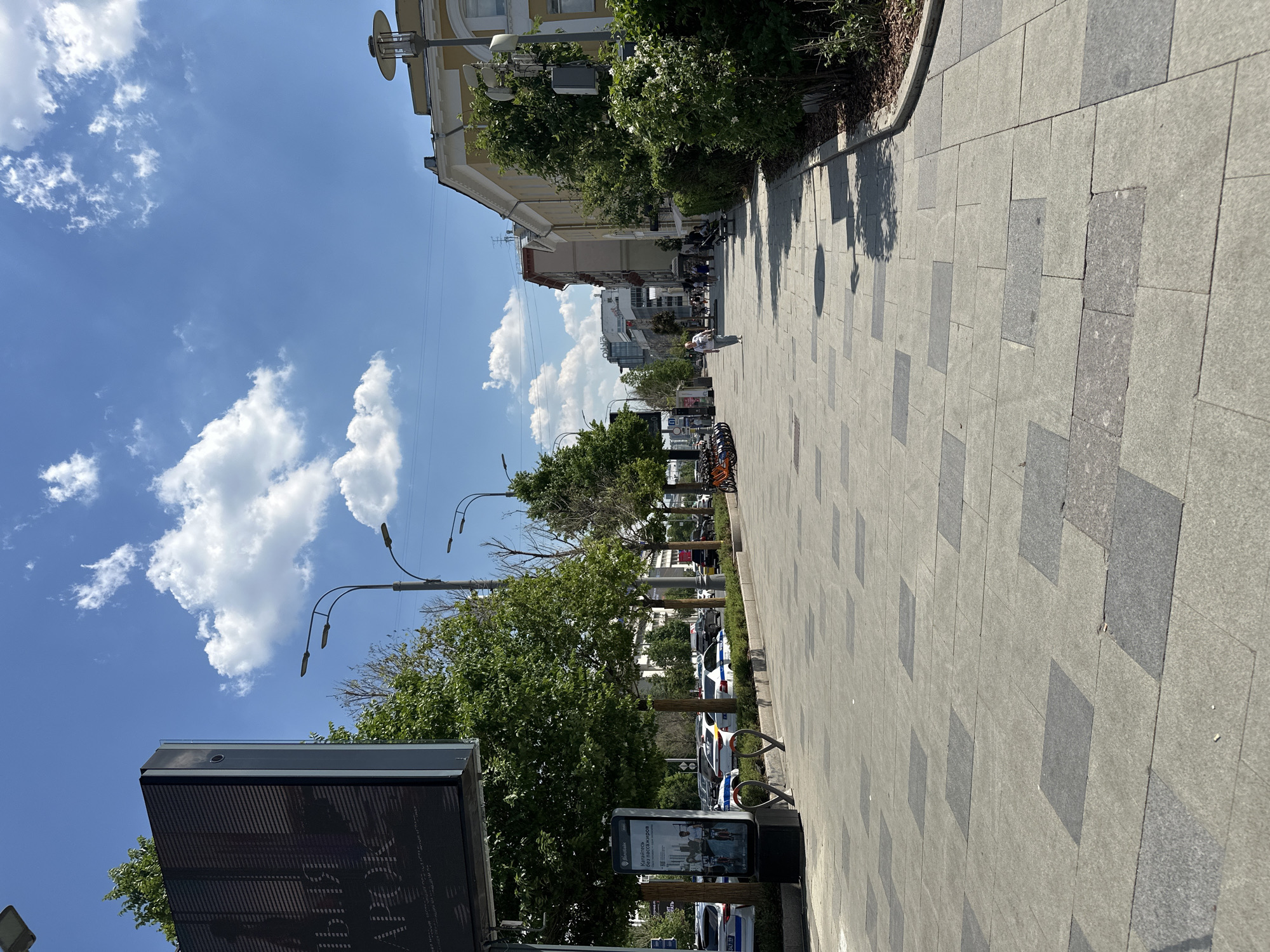 The image captures a sunny day on a wide pedestrian walkway in Moscow, Russia. The walkway is paved with light grey and dark grey rectangular tiles arranged in a patterned grid. On the left side of the walkway, a large digital billboard displays text in Russian. Adjacent to the billboard, there are bike racks with several orange bicycles parked in them. A digital display board is also visible near the bike racks.
Lined along the walkway are several mature trees with vibrant green foliage, casting dappled shadows on the pavement. Beyond the trees, cars are parked along a street. In the distance, various multi-story buildings are visible, some with light-colored facades and others with more modern glass exteriors.
A woman wearing a light-colored top and dark trousers is walking along the walkway, away from the camera, with a backpack. Several other pedestrians are further down the walkway, some sitting on benches and others walking. Streetlights with a modern design are positioned along the walkway and street. The sky is a bright blue with scattered white cumulus clouds. The overall scene depicts a well-maintained urban public space on a clear day.
