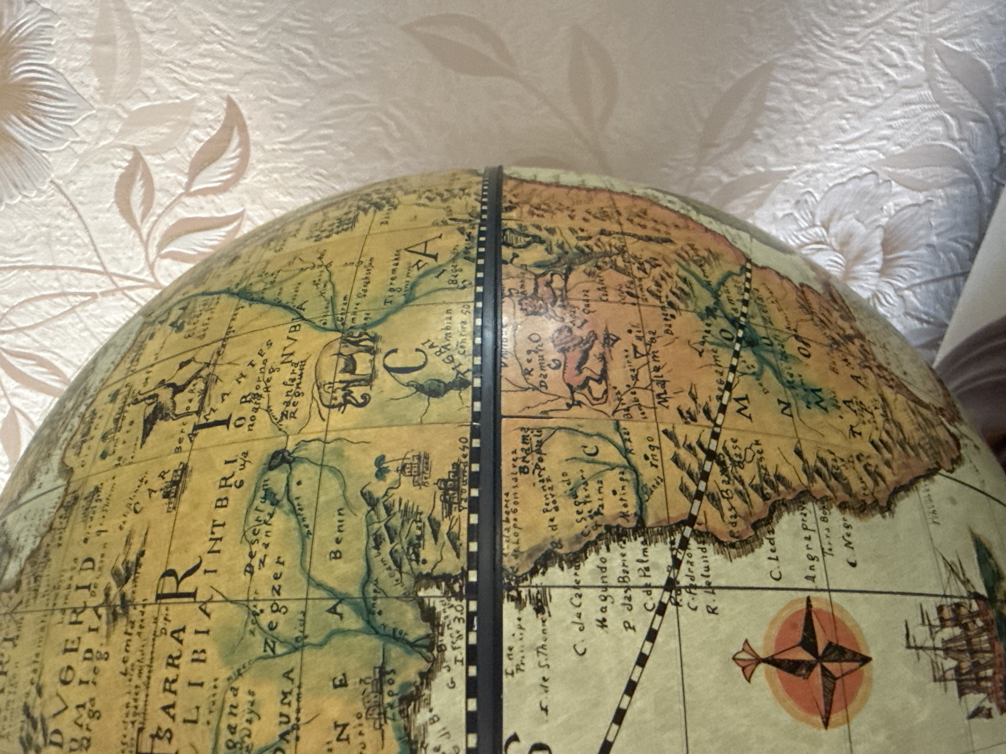 The image depicts a close-up view of an antique globe, showcasing a portion of Africa. Place names such as "LIBIA," "Benin," and "Negra" are visible, along with geographical features like "Desertum Zegzer." The map style appears historical, with detailed drawings and cursive lettering. A dark vertical strip divides the globe, indicating a seam in its construction. A compass rose and a depiction of a sailing ship are present on the lower right section. The background is blurred and appears to be a patterned wall or fabric. There's no indication of Адам, Russia.