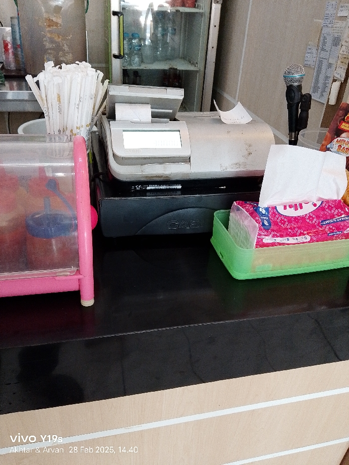 The image shows a cash register and other objects on a counter in a shop. The shop appears to be in a city, based on the surroundings and objects in the image. The cash register is on the counter and is black and silver in color. There are a variety of other objects on the counter as well, including a pink plastic container, a stack of white paper straws, and a green plastic container with a pink and white tissue box. There is also a microphone on the counter. The counter is black and has a white wood surface underneath. The overall scene suggests that the shop is likely a food or beverage establishment, given the presence of a cash register, straws, and napkins. The image is taken from a slightly elevated angle, with the counter being in the foreground. The location is a shop in Patrol, Indonesia, with coordinates -6.318 latitude and 107.986 longitude.