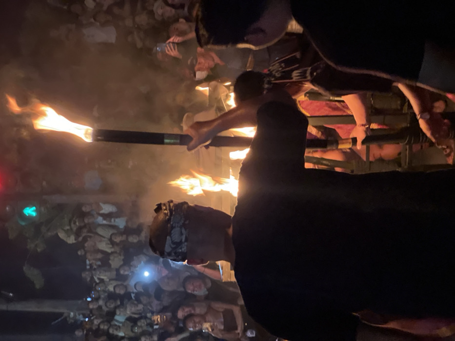The image captures an outdoor night scene in Munggu, Indonesia, during a torchlit event. In the foreground, a person seen from behind wears a black short-sleeved shirt and a patterned head covering, holding a tall black torch with an upward-streaming orange flame in their right hand. Multiple other torches with bright flames are visible, generating significant smoke that diffuses the background. To the right of the central figure, a second individual is partially visible in dark clothing. Further right and slightly lower, a third person in red or maroon attire, wearing a distinct headdress, is also interacting with a torch or a fiery element. A dense crowd of spectators extends into the background, with many individuals holding up smartphones, some with active flashlights, to record the event. Overhead, a traffic light displays a green pedestrian signal and a red vehicle signal.