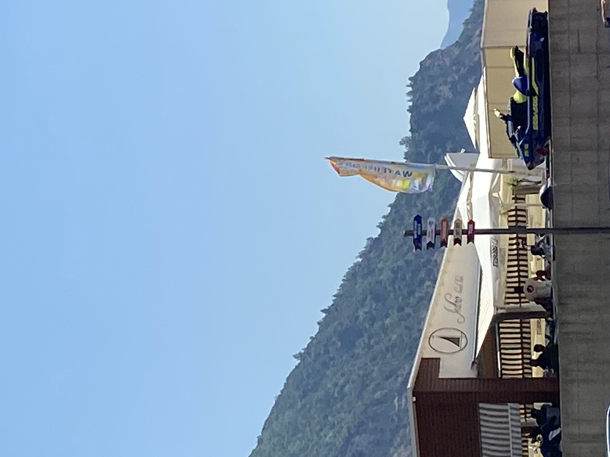 The image shows a clear blue sky above a tree-covered mountainside. In the foreground, a building with a brown facade and a striped awning is visible. The building appears to be a cafe or restaurant, with signage that reads "Jadran". Several people are seated on a low wall in front of the building. A jet ski is parked on the wall to the right of the building. A flag with an orange and yellow design is flying from a pole behind the building. Traffic signs are also visible on a pole to the right of the building, pointing in different directions. The overall scene suggests a sunny day at a waterfront establishment.