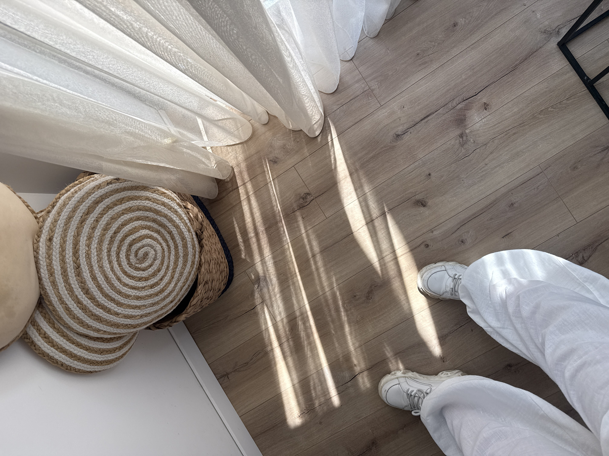 An eye-level, interior shot shows a person in white pants and white sneakers standing on light brown wood flooring, with the light streaming from a sheer, white curtain in the upper left. To the left is a round, woven storage basket with white and tan spiral bands. The location is an interior in Varna, Bulgaria.