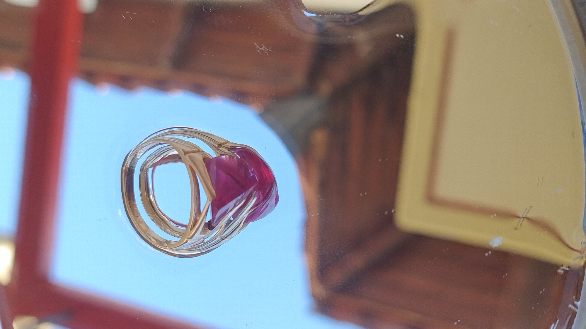 A gold ring with a faceted, deep pink gemstone is reflected on a shiny, transparent surface. The ring appears to have a "14K" stamp visible on the band. The reflection shows the ring suspended against a bright blue sky. In the blurred foreground and background, there are reflections of brown and yellow structures, possibly buildings or furniture. The environment is indoor, with the ring placed on a reflective surface. No people are present in the scene.