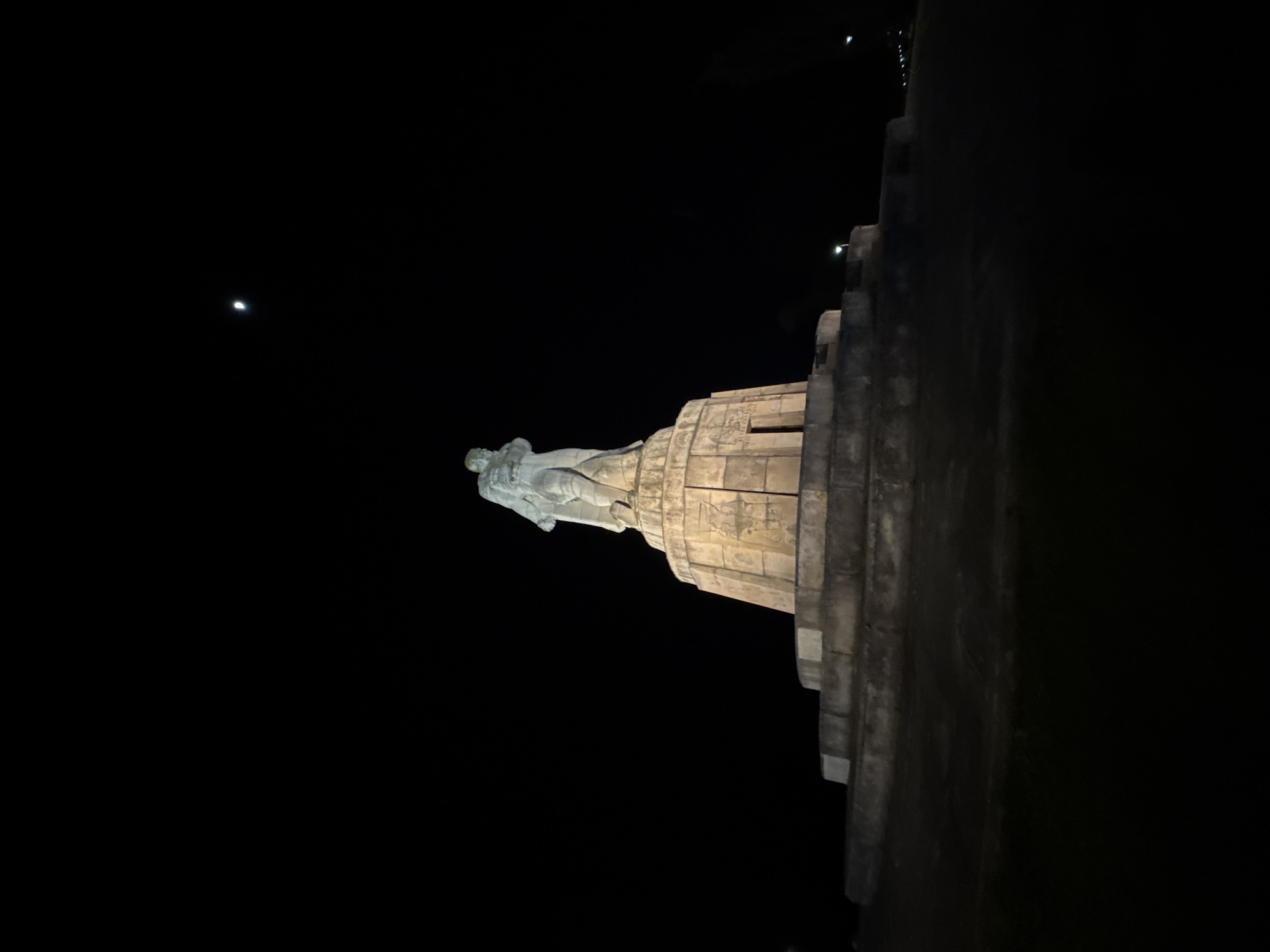The image displays a large, illuminated stone monument captured at night, set against a deep black sky with a visible crescent moon. The monument consists of a prominent, heroic-style human figure, likely male, standing atop a substantial, tiered architectural pedestal. Both the statue and its base are made of a light-colored material, possibly concrete or stone, and are brightly lit by artificial lights, creating a stark contrast with the dark surroundings.
The figure of the statue appears to be in a resolute or commanding pose, with the details of its form highlighted by the lighting. The multi-layered pedestal exhibits carved or relief elements on its surfaces, though no specific text is legible. In the upper left quadrant of the dark sky, a small, distinct crescent moon is visible, indicating clear weather. Faint, distant lights can also be discerned on the far right edge of the frame, possibly from nearby structures or city lights. The scene evokes a sense of solemnity and historical grandeur. Given the context of Varna, Bulgaria, this is most likely the Monument of the Bulgarian-Soviet Friendship.