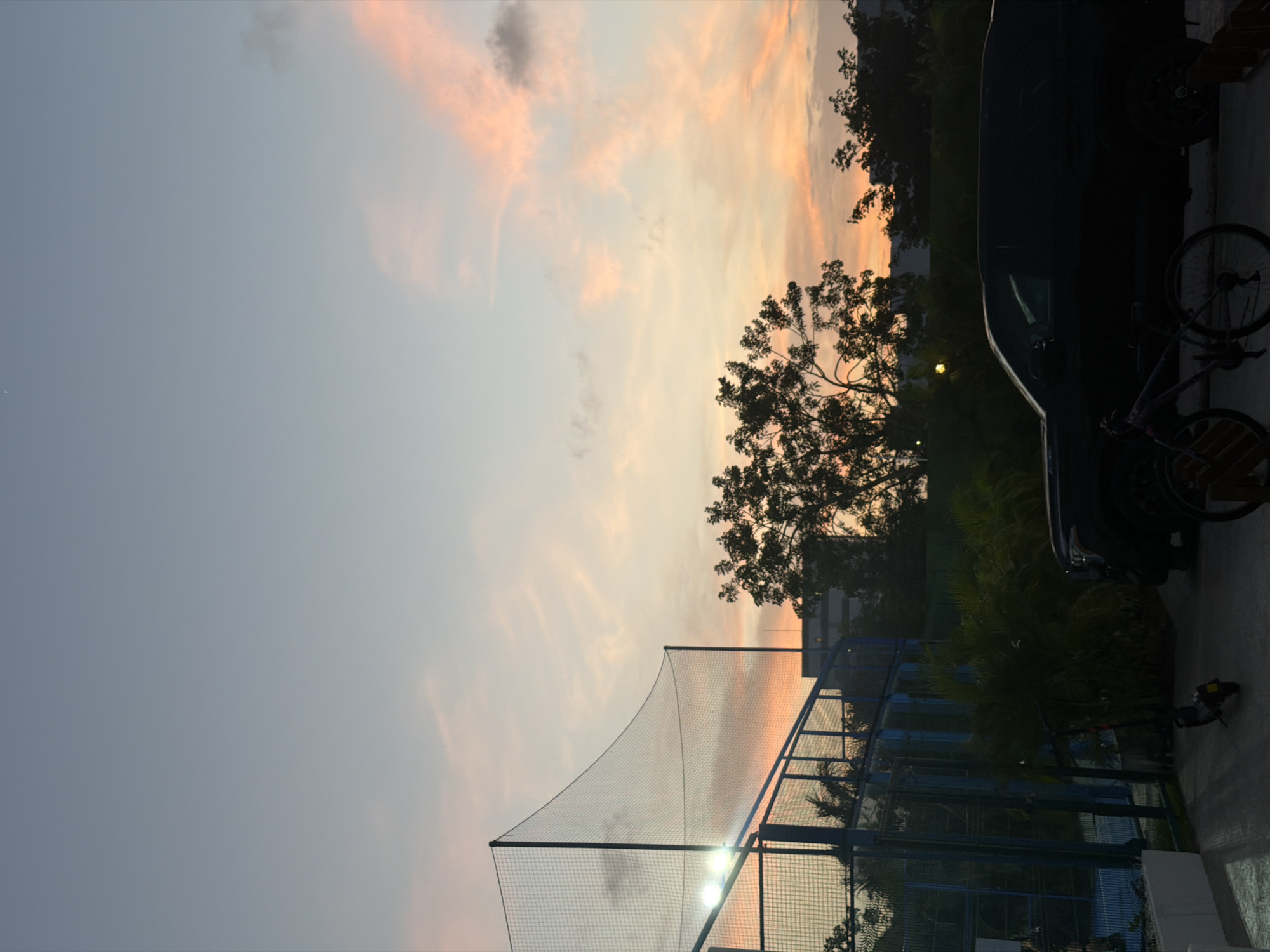 The image captures an outdoor scene at sunset in Cancún, Mexico, showcasing a serene sky transitioning from a soft blue-grey at the top left to warm orange and pink hues towards the horizon on the right. Scattered, wispy clouds are visible across the sky.
In the foreground and midground, silhouetted trees with dense foliage are prominent, particularly on the right side and lower center, indicating a tropical or subtropical environment. A tall, light-colored netting, possibly belonging to a sports facility such as a batting cage or tennis court, stretches from the lower left towards the center and right, supported by vertical poles. Through and behind this netting, a blue-framed fence runs parallel.
On the far right, a dark vehicle, likely a car or van, is parked and largely enveloped in shadow. A bicycle leans against or near this vehicle. A bright artificial light source, possibly a floodlight, shines through the netting on the lower right, and another fainter light is visible further in the background. Distant buildings or structures are dimly discernible through the trees and netting, suggesting an urbanized area.
No specific activity is taking place; the scene is a tranquil snapshot of dusk. The warm colors of the sky evoke a calm and peaceful atmosphere. There is no visible text in the image.