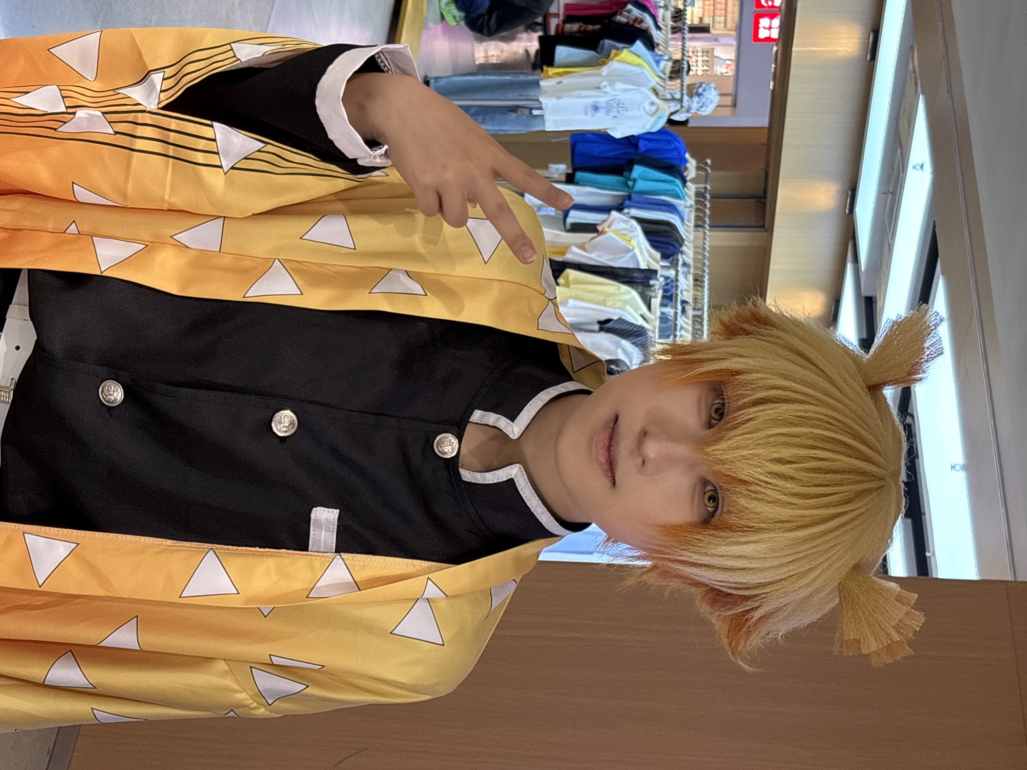 A person dressed in a yellow and black costume with a blonde wig, possibly a character from an anime or manga series, is seen making a peace sign with their fingers. The background appears to be a retail environment, with clothing racks and shelves stocked with various items. A storefront with Chinese writing on a sign is visible in the distance. This photograph was taken in the city of Liangjiang, China.
