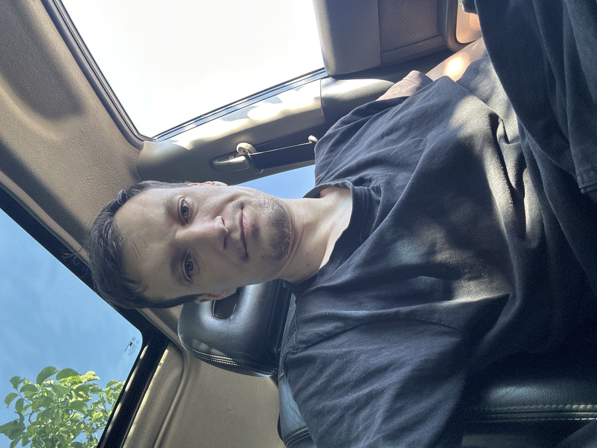 A man is sitting in the driver's seat of a car, taking a selfie. He is wearing a black t-shirt. The car has a sunroof, through which a blue sky and green leaves can be seen. The man's face is partially obscured by the angle of the photo, but his eyes, nose, and mouth are visible. He appears to have short, dark hair and is clean-shaven with a faint stubble. The car's interior, including the leather seat and dashboard, is visible. The image is taken during the daytime, as indicated by the bright sunlight streaming through the sunroof and windows. There is no information in the image to suggest the location is Shatava, Ukraine.