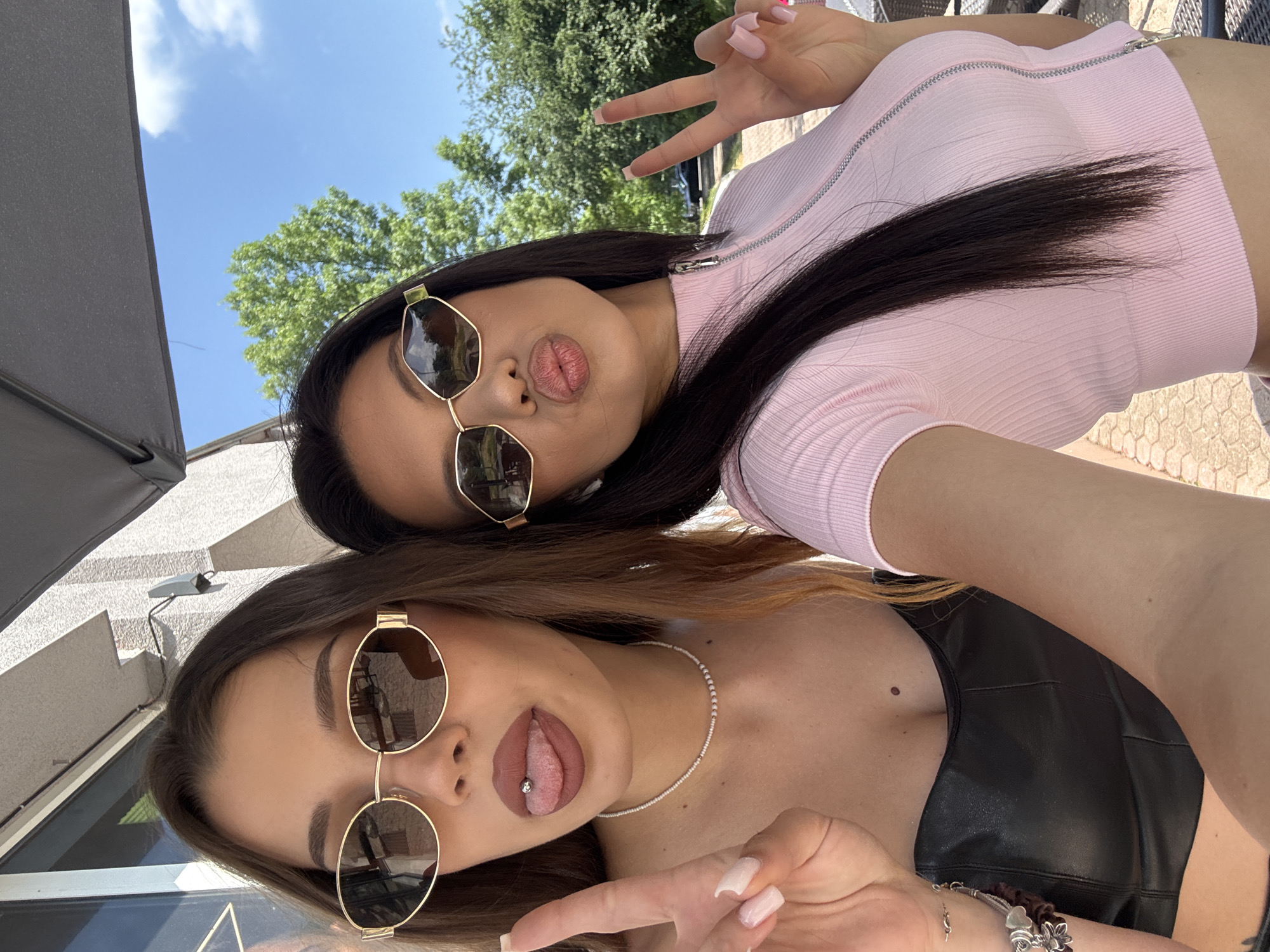 Two young women are captured in a playful outdoor selfie, likely in Sofia, Bulgaria, judging by the provided context. The image is rotated counter-clockwise.
The woman positioned higher in the frame has long, dark hair and is wearing a light pink ribbed crop top with a half-zip front. She sports gold-framed hexagonal sunglasses and makes a peace sign with her right hand, while her lips are puckered into a 'duck face'. Her nails are long and painted light pink.
The woman in the foreground has brown hair and is wearing a black, leather-look strapless top. She wears gold-framed round sunglasses and has her tongue playfully sticking out, revealing a tongue piercing. A delicate white beaded necklace adorns her neck, and a silver charm bracelet is visible on her wrist. She also has light pink acrylic nails and appears to be making a peace sign with her left hand, with only parts of her fingers visible.
The setting is outdoors on a bright, sunny day, indicated by the clear blue sky with some scattered white clouds and lush green trees in the background. On the left side, a dark grey umbrella or awning is partially visible. Behind the foreground woman, a light-colored building with some architectural features can be seen. The ground beneath them appears to be paved with light-colored bricks or tiles. Both women convey a relaxed and cheerful mood. No visible text is present in the image.