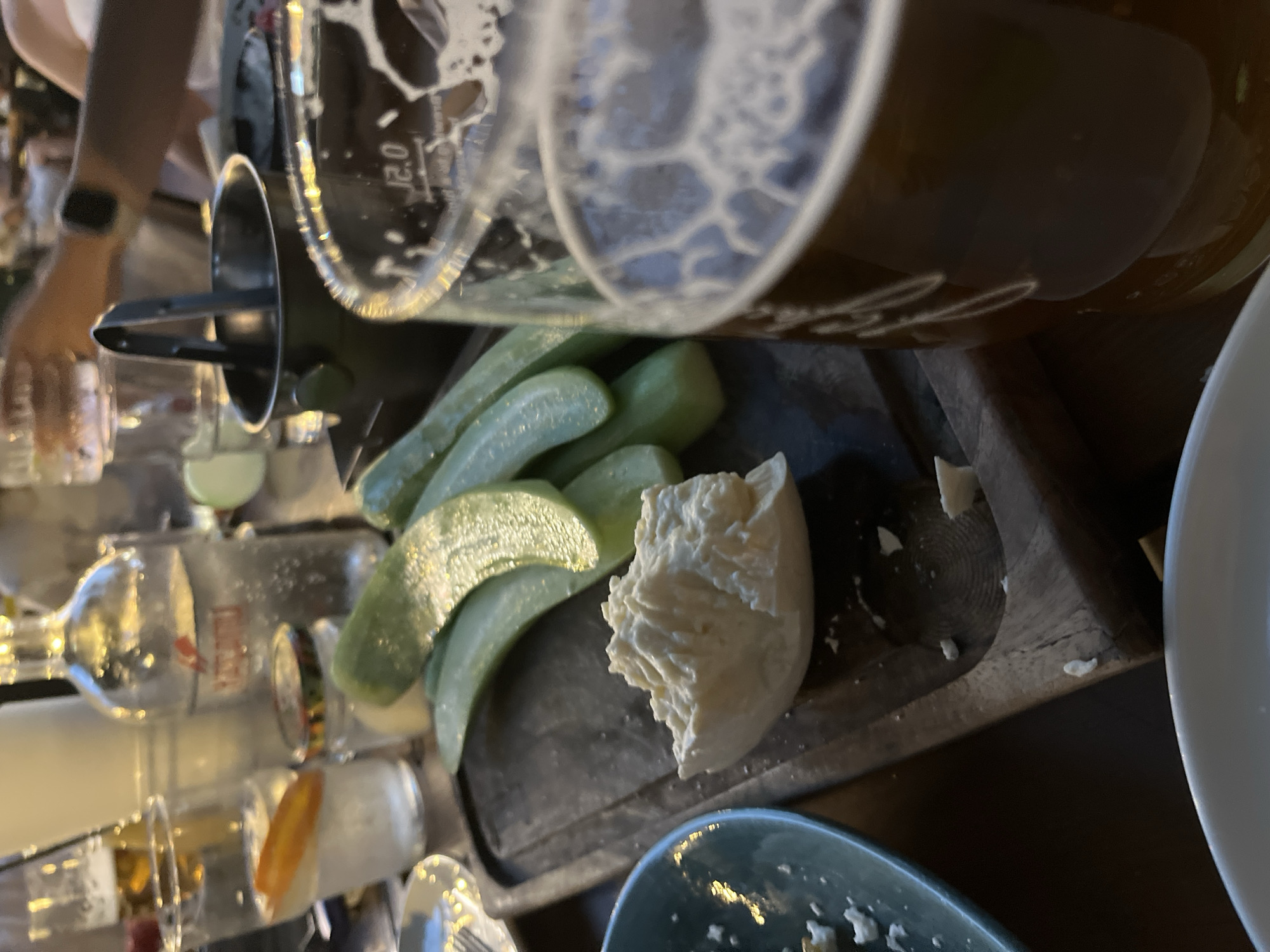 This image depicts a table scene in a restaurant or bar located in Plovdiv, Bulgaria, captured at close range with a shallow depth of field. In the immediate foreground, to the left, a light blue ceramic bowl containing scattered white crumbs is partially visible. Centrally, on a dark wooden serving board, rests a substantial piece of white, crumbly cheese, likely feta, accompanied by several green, elongated slices of cucumber. To the right, a clear glass holding a dark beverage, presumably beer, with a foamy head, displays "12.0" markings and is slightly out of focus.
In the mid-ground, behind the central board and beer glass, an assortment of drinks and accessories are present. These include a small, clear glass jar with a lid, a tall, clear glass bottle containing a carbonated clear liquid and featuring a red logo and partially legible text, a dark metal ice bucket holding black tongs, and another clear drinking glass containing a slice of orange or lemon in a clear liquid. The background is blurred, showing indistinct figures of other patrons and general restaurant lighting. A human hand and forearm are discernible in the upper right background, suggesting activity. The overall lighting is dim and warm, indicative of an evening dining atmosphere.