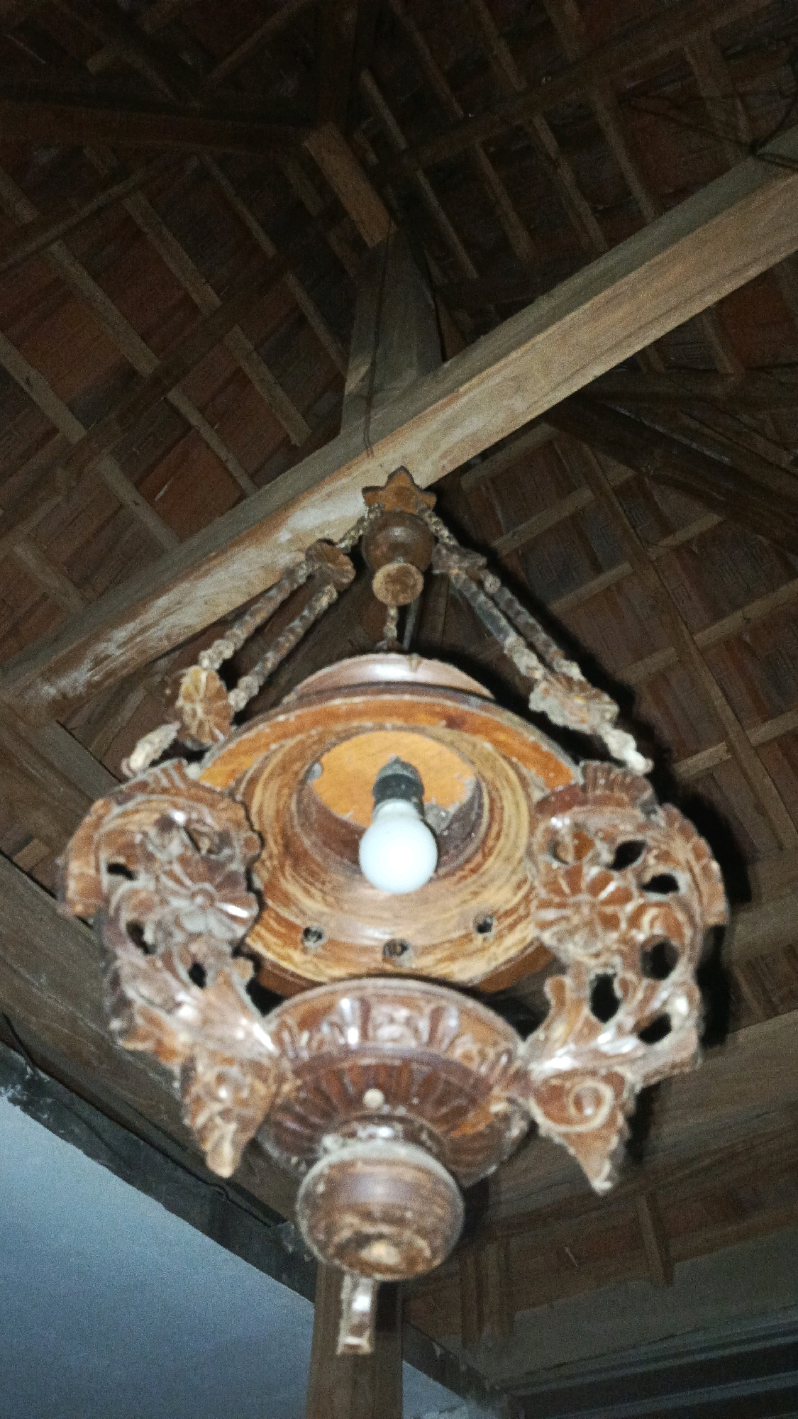 A carved wooden chandelier hangs from a wooden ceiling structure in what appears to be an interior space. The chandelier is intricately detailed with floral and possibly animalistic motifs. A single light bulb is visible within the central part of the fixture. The ceiling is composed of exposed wooden beams and what appear to be terracotta tiles. The overall impression is of an older, possibly traditional, architectural style. The specific location in Babadan, Indonesia, is inferred from the context provided, though not visually evident from the image itself.
