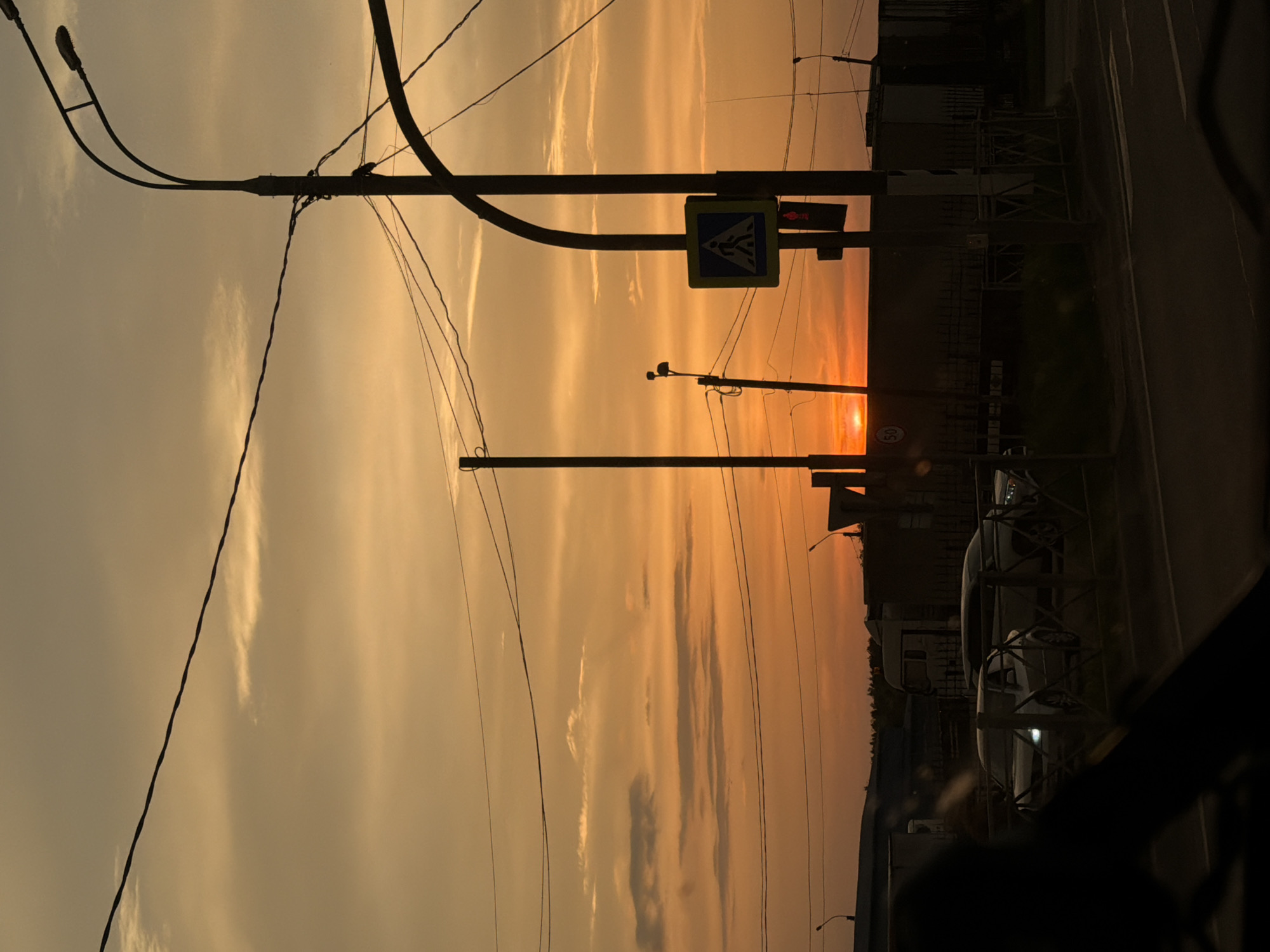 The image captures a sunset in Спас-Заулок, Russia. The sky is a gradient of yellow and orange hues, with streaks of clouds. Silhouetted against the sky are various objects: utility poles with wires, a pedestrian crossing sign, a streetlight, and buildings. On the right side of the image is a road and partially visible vehicles behind a fence. The lower right corner is dark, indicating the vantage point is from inside a vehicle.