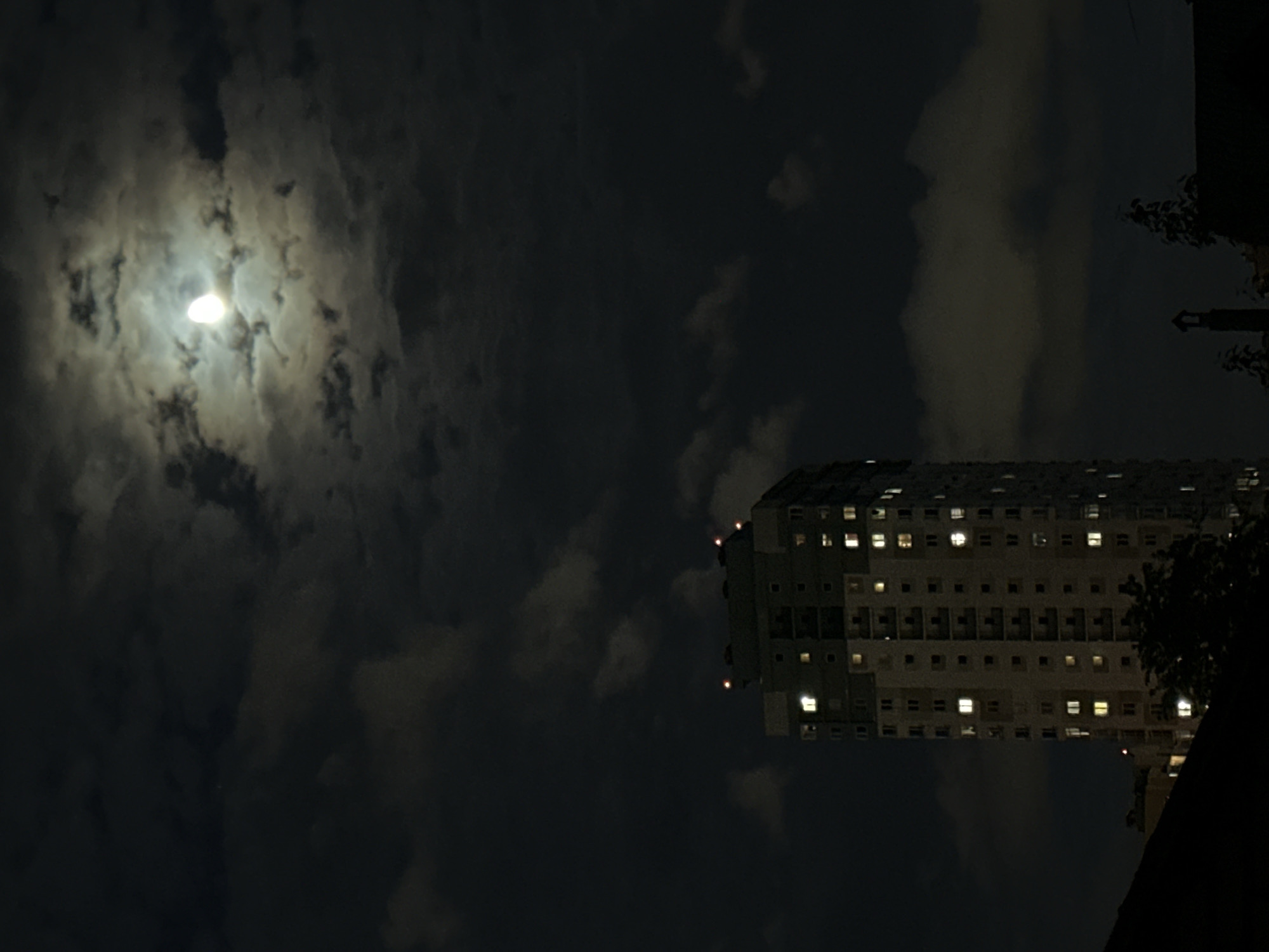 The image shows a night scene with a bright moon partially obscured by clouds in the upper left quadrant. To the right is a tall, rectangular building with many lit windows, indicating it's likely residential. The building appears to be in Minsk, Belarus, given that the image originates from there. Dark trees and foliage are present in the lower right corner, suggesting a park or green space in the vicinity. The overall scene appears to be urban, captured at night.
