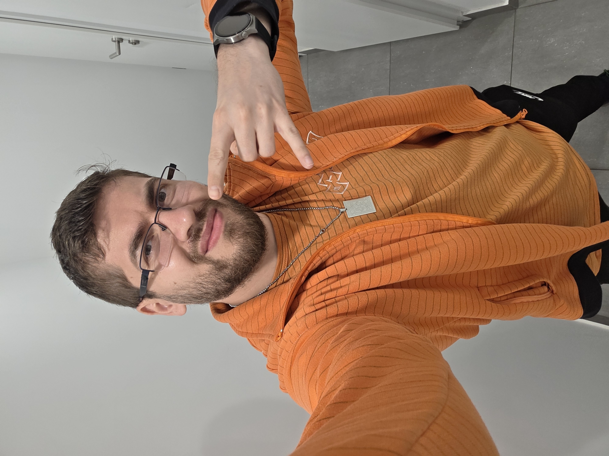 A man is taking a selfie indoors. He has brown hair and a beard, wears glasses, an orange striped jacket, and a watch. He is pointing towards his face with his right hand. He wears a chain necklace with a silver tag. The background includes a white wall and gray tiles. It is impossible to determine if this photo was taken in Plovdiv, Bulgaria, based on the visual information provided.