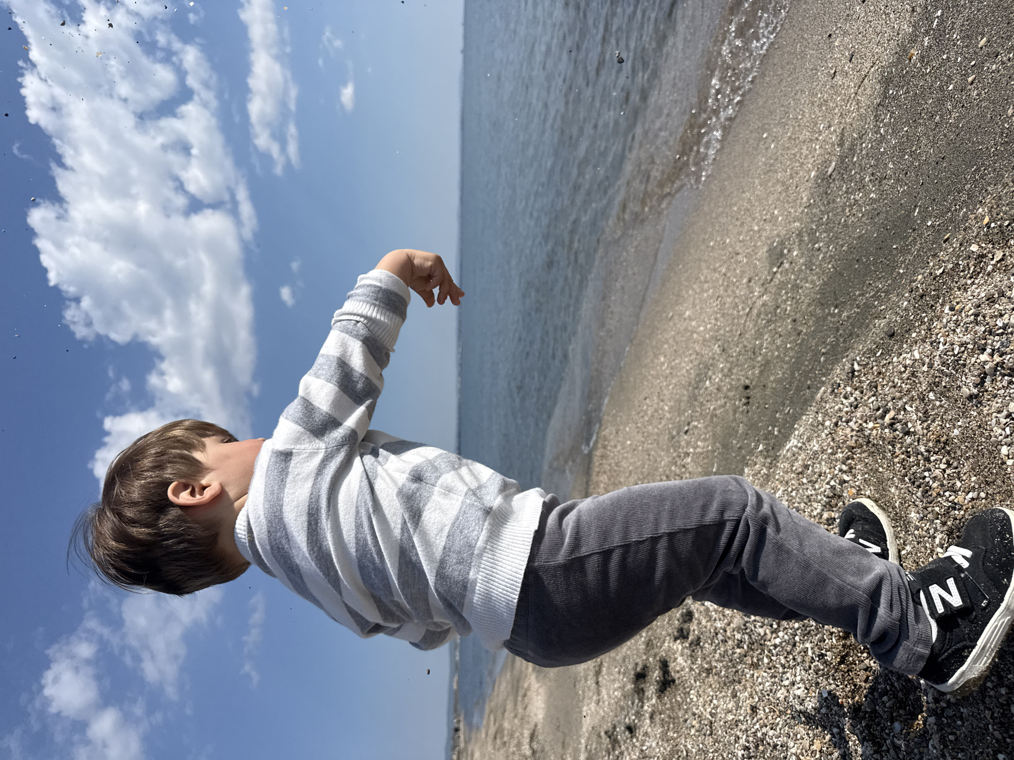 A child stands on a beach. The beach consists of sand and small pebbles. The sea laps the shore. The sky is partly cloudy. The child wears a striped sweater, gray pants, and black sneakers with a white "N" logo. The child's hand is outstretched towards the water. The photograph was taken at a tilted angle. Location: Likely a beach in Burgas, Bulgaria.