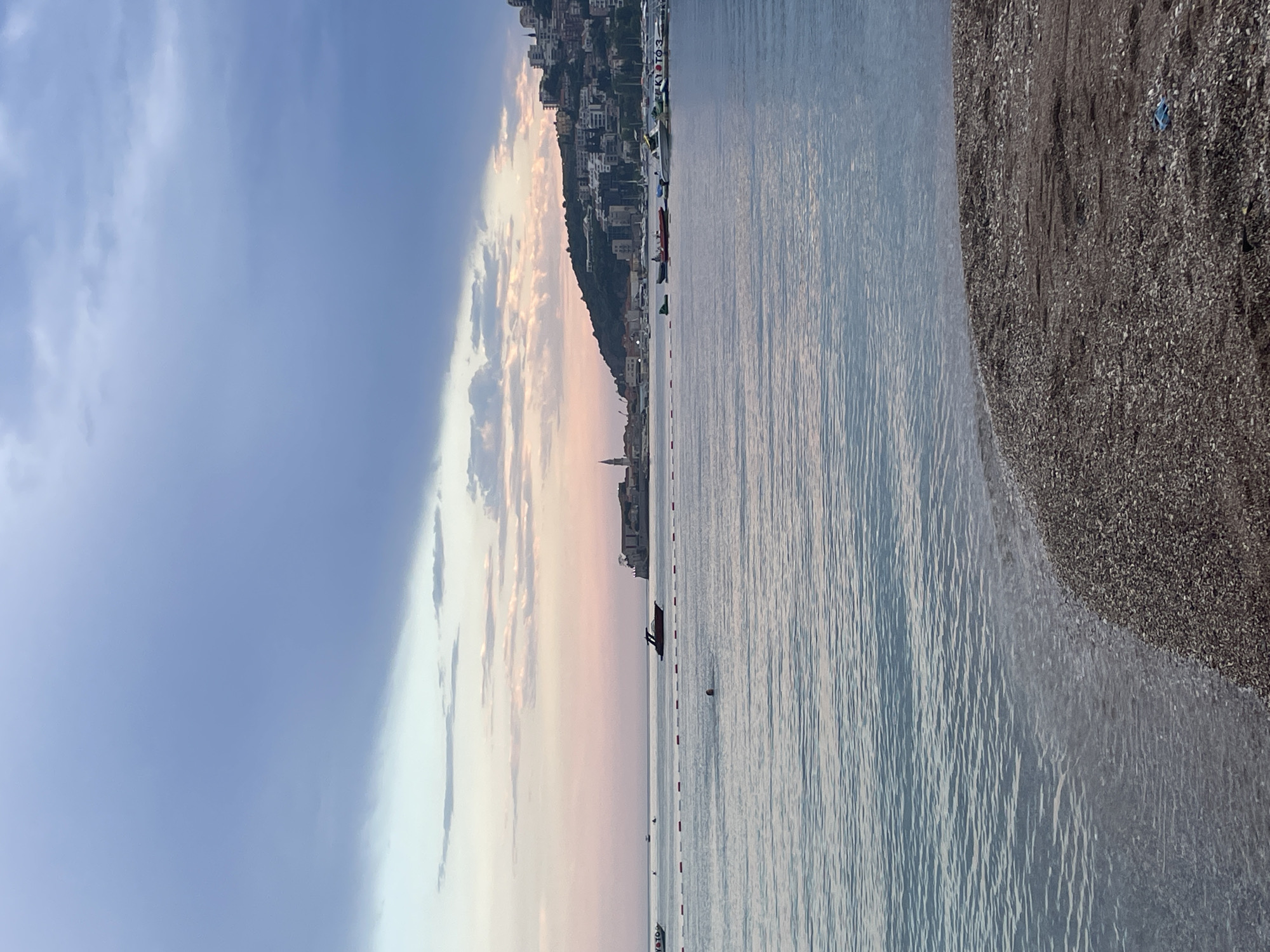This is a landscape photograph taken at the beach in Budva, Montenegro. The image is oriented vertically. The left side of the frame shows a dramatic sky with a mix of dark clouds and areas of soft, pastel-colored clouds, suggesting either dawn or dusk. A city built on a hillside is visible in the distance, with buildings cascading down towards the water. The sea occupies the right two-thirds of the image, appearing calm with gentle ripples reflecting the sky's colors. A shoreline of dark pebbles is visible on the far right.
Several objects are scattered across the water, including what appear to be buoys or markers, and a few small boats. In the mid-ground, on the water, a small structure with what looks like a red roof is visible, possibly a small floating platform or a boat. There are also some people visible in the water, some on the platform, and a few dots that could be swimmers further out. The overall mood is serene and peaceful, enhanced by the soft light and the quiet atmosphere of the beach at what is likely the end of the day. A small spire or tower is visible among the buildings on the hillside. Some faint text is visible on a boat or structure in the water that reads "KOTOB3".
