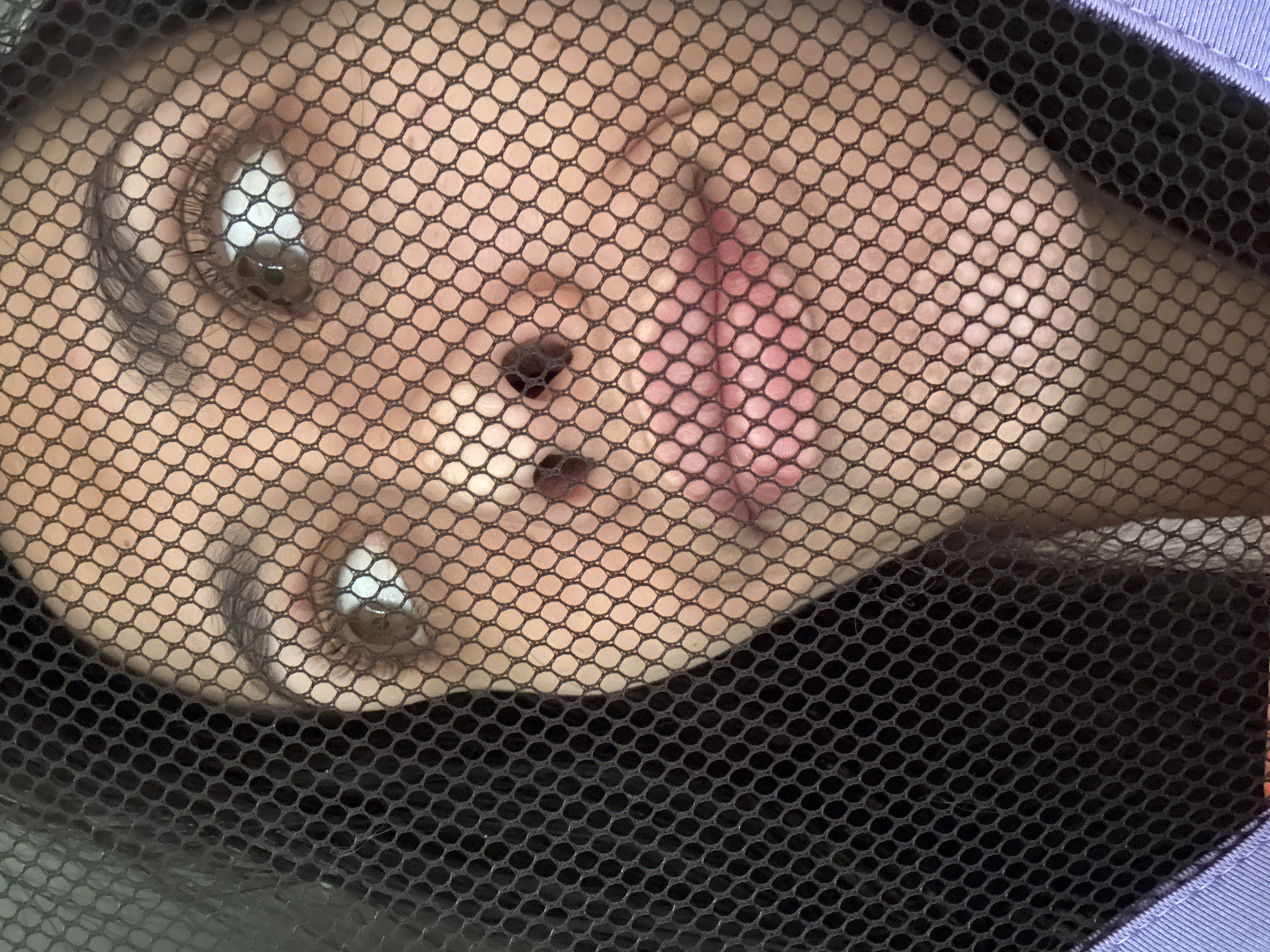 A close-up, overhead shot shows a woman looking upwards through a black mesh fabric. Her face is partially obscured by the mesh, creating a pattern of circles over her features. The woman has dark hair, visible on the left side of the frame, and appears to be wearing light-colored clothing, with a hint of purple fabric visible at the bottom edge of the image. The background is dark and indistinct. There is no discernible location within the city of Votkinsk, Russia.
