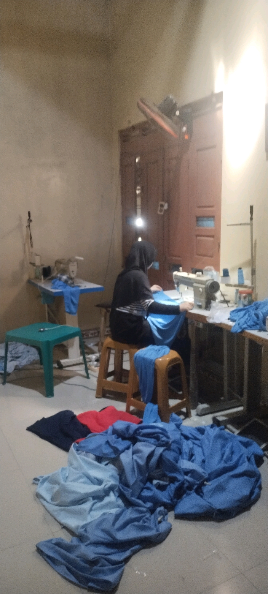 A wide shot captures an interior scene in a workshop or tailoring establishment, likely located in Daleman, Indonesia. The room is simply furnished with a concrete floor and beige walls. In the foreground, a pile of fabric, predominantly in shades of blue with some red and dark blue pieces, lies on the floor. To the left, a small, turquoise plastic table stands with a sewing machine on it. Further back, a person wearing a dark headscarf and a striped shirt is seated on a wooden stool, actively engaged in operating a sewing machine. The machine is situated on a table to the right of the seated individual. Behind the person and the sewing machine, a set of closed wooden doors is visible, with a bright light source illuminating the upper portion of the frame, suggesting a window or overhead lighting. The overall impression is one of a functional, working space dedicated to textile production.