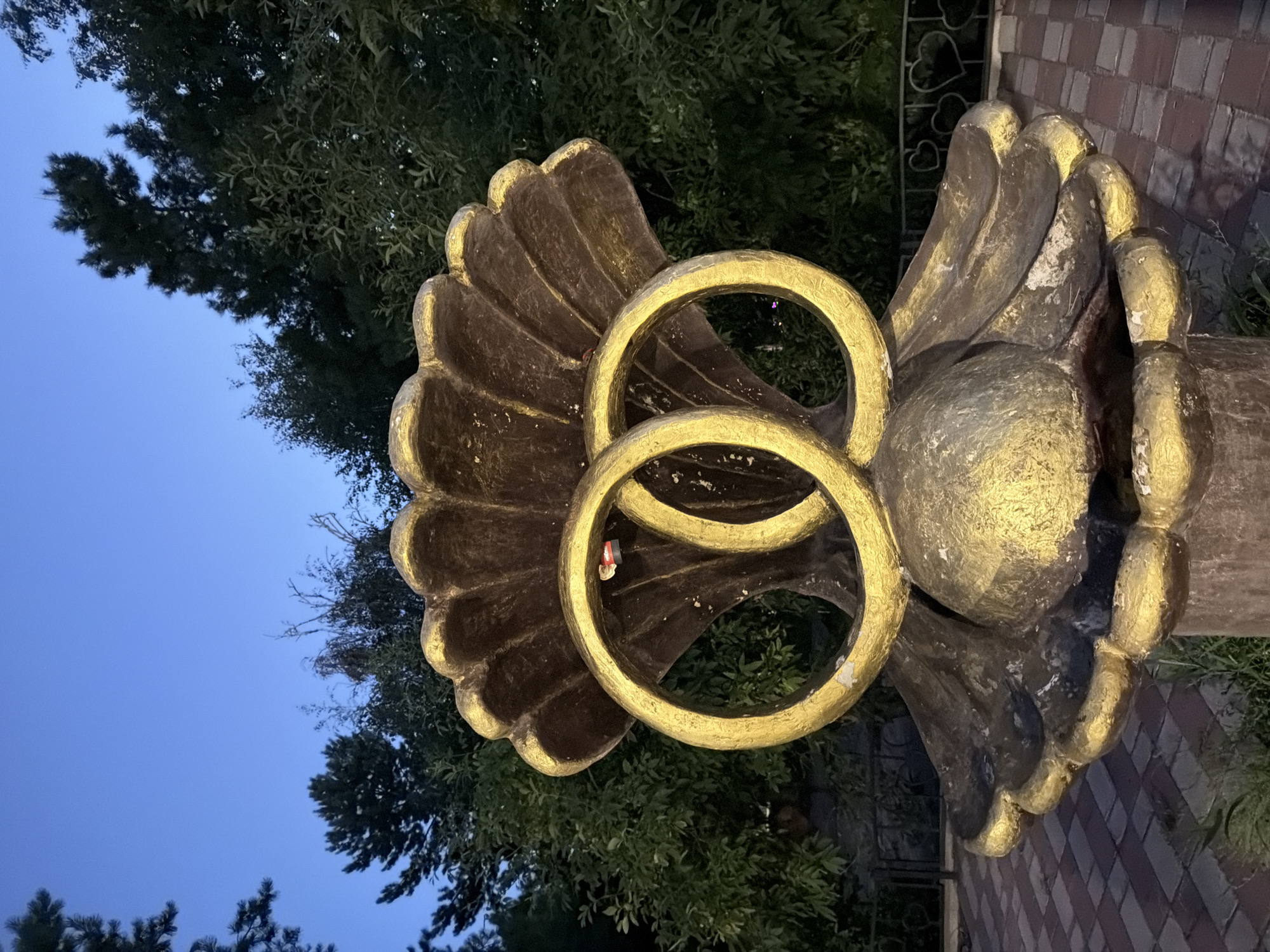 An outdoor sculpture of two interlocked gold rings is featured prominently in the center of the image. The sculpture appears to be made of metal and is weathered, with some areas of rust and chipping paint. It is situated in front of a dense background of green trees and foliage. The sky above is a deep blue, suggesting it is either dusk or dawn. In the foreground, a paved area with a brick-like pattern is visible. A black metal fence with heart-shaped designs can be seen behind the sculpture. The overall impression is that of a decorative element in a park or public space, likely a monument related to marriage or commitment. This sculpture is located in the city of Pavlodar, Kazakhstan.