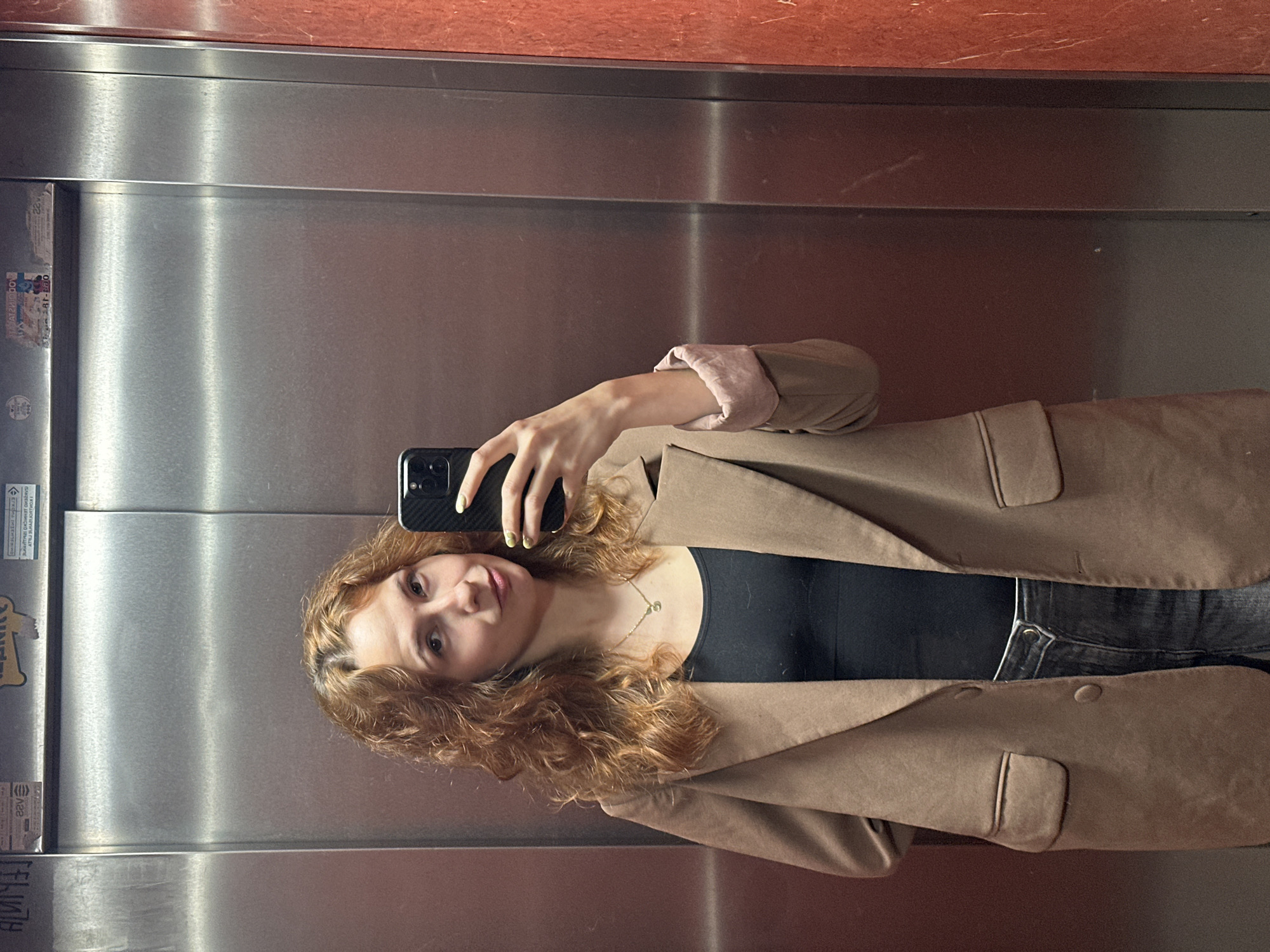 A woman is taking a selfie in an elevator with a stainless steel interior in Novi Sad, Serbia. The upper portion of the elevator walls is covered with a red marble-like material. The woman has curly red-blonde hair and is wearing a black top and a brown blazer. She holds a black smartphone with a black case in her right hand. Her fingernails are painted with pale yellow nail polish. A thin gold necklace is visible around her neck. The elevator has informational stickers and labels on the left wall.