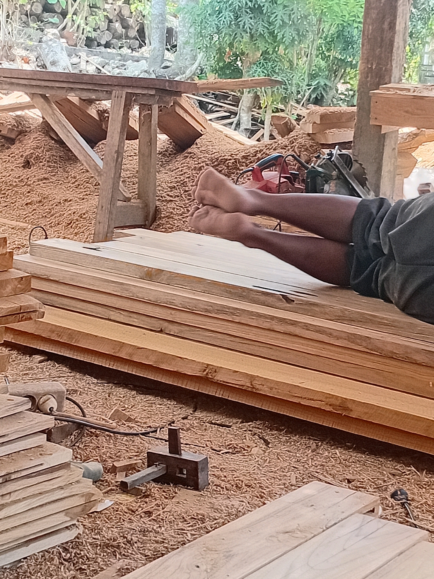 In Playen, Indonesia, a person is seen lying down with their legs extended across a stack of lumber. The environment appears to be a woodworking area, characterized by piles of wood shavings on the ground. Various tools and materials related to woodworking are visible, including what appears to be a circular saw, a power tool, and other wooden planks. The scene suggests a workspace where lumber is being processed or stored.