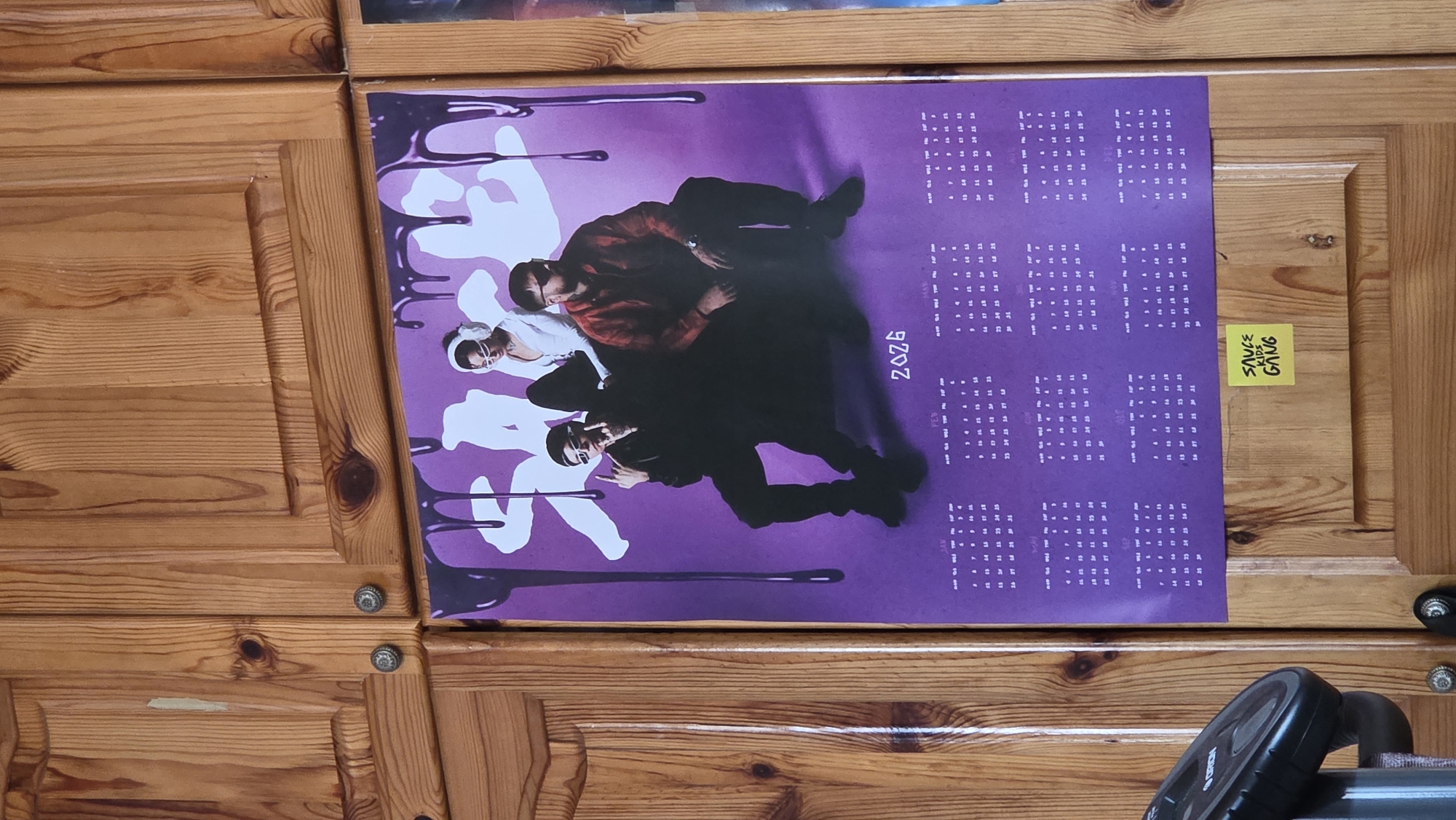 The image shows a poster taped to a wooden cabinet. The poster is purple and features three people stacked on top of each other, with white and purple designs resembling drips. The poster also has a calendar grid labeled "2026". A yellow sticky note with "SAUCE KIDS GANG" written on it is attached next to the poster. A black object, possibly part of a machine, is partially visible at the bottom right corner. The background is the wooden cabinet with visible grain and knobs. The location and city are unknown.
