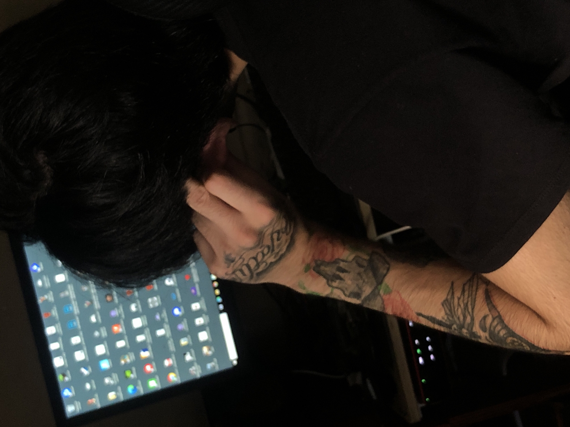 A person with dark hair is seated in front of a computer monitor displaying numerous application icons. The person has tattoos on their forearm and hand. The location in Buenos Aires, Argentina is not discernible from the image. The person appears to be adjusting or touching their ear with their hand. The person is wearing a black garment. The environment is dimly lit.